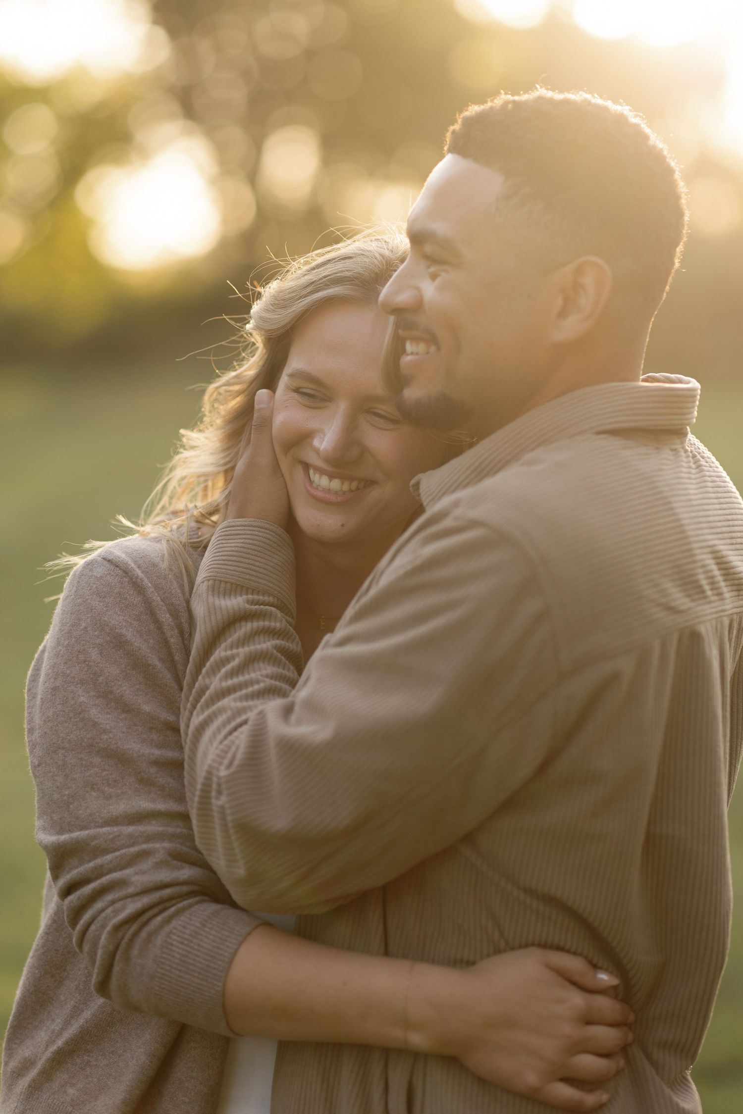 A couple embraces warmly in an outdoor setting during golden hour with soft bokeh lighting in the background.
