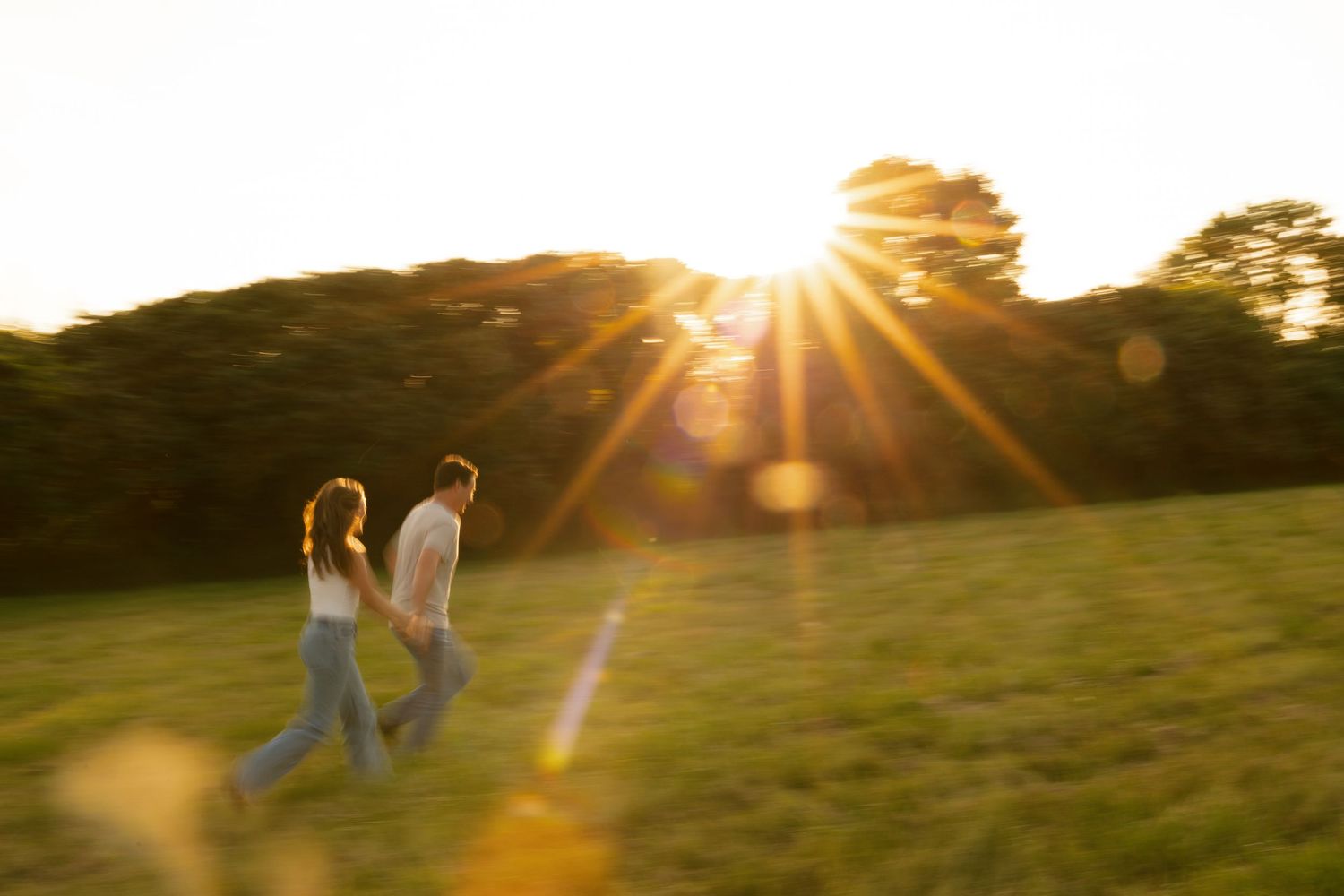 People walking on a grassy hillside at sunset with golden sunlight streaming through trees in the background.