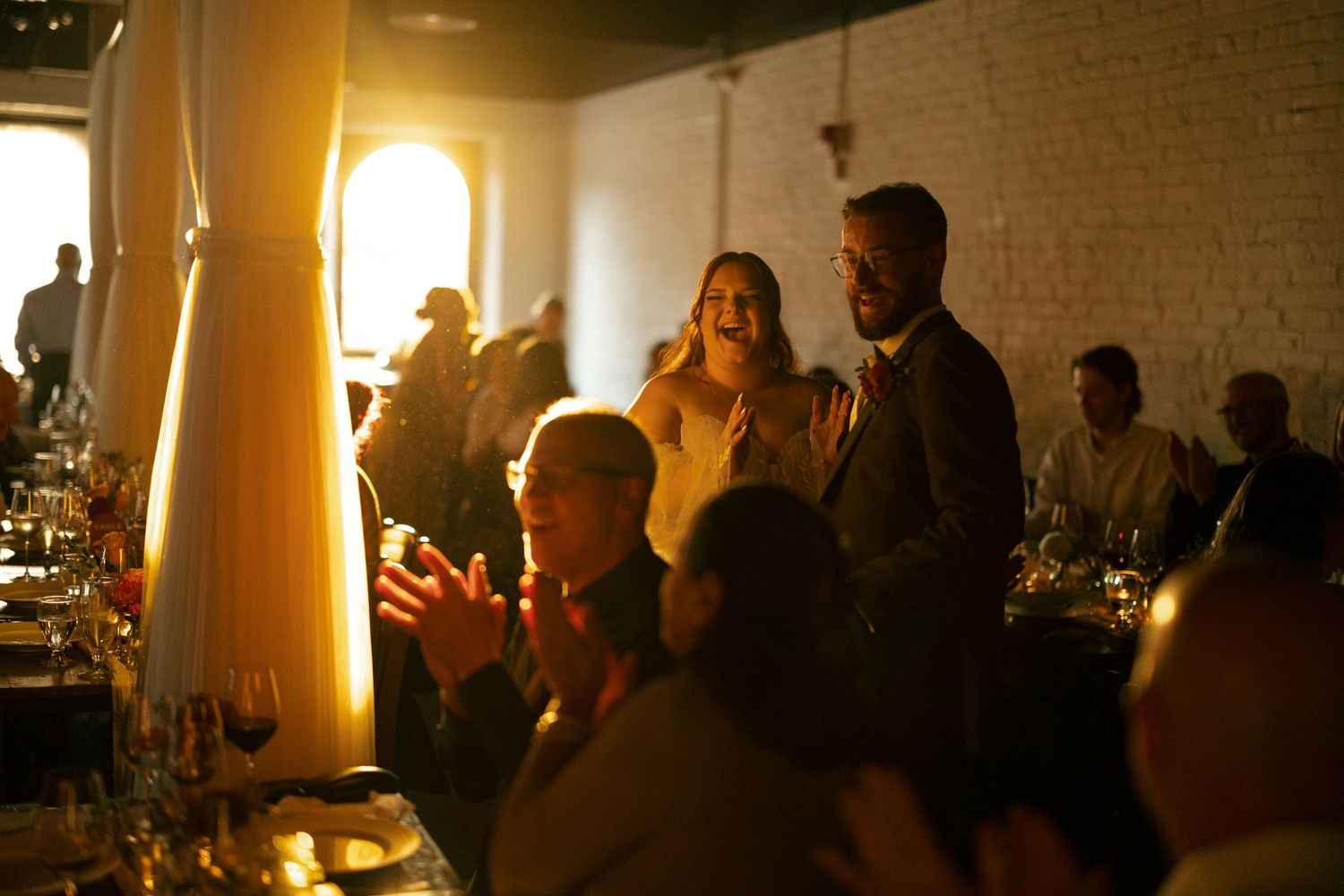 People gather in a warmly lit indoor venue with brick walls and white columns during an evening event.