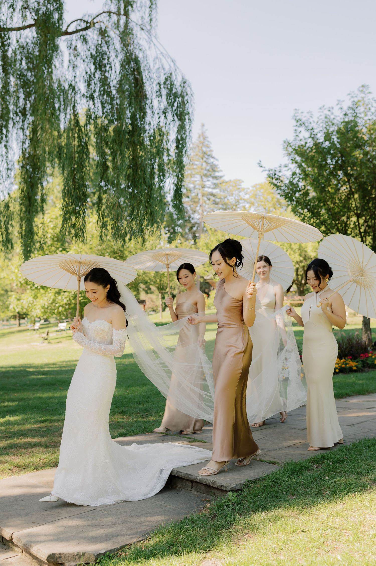 A group in light colored dresses and parasols pose playfully in a garden setting on a sunny day.