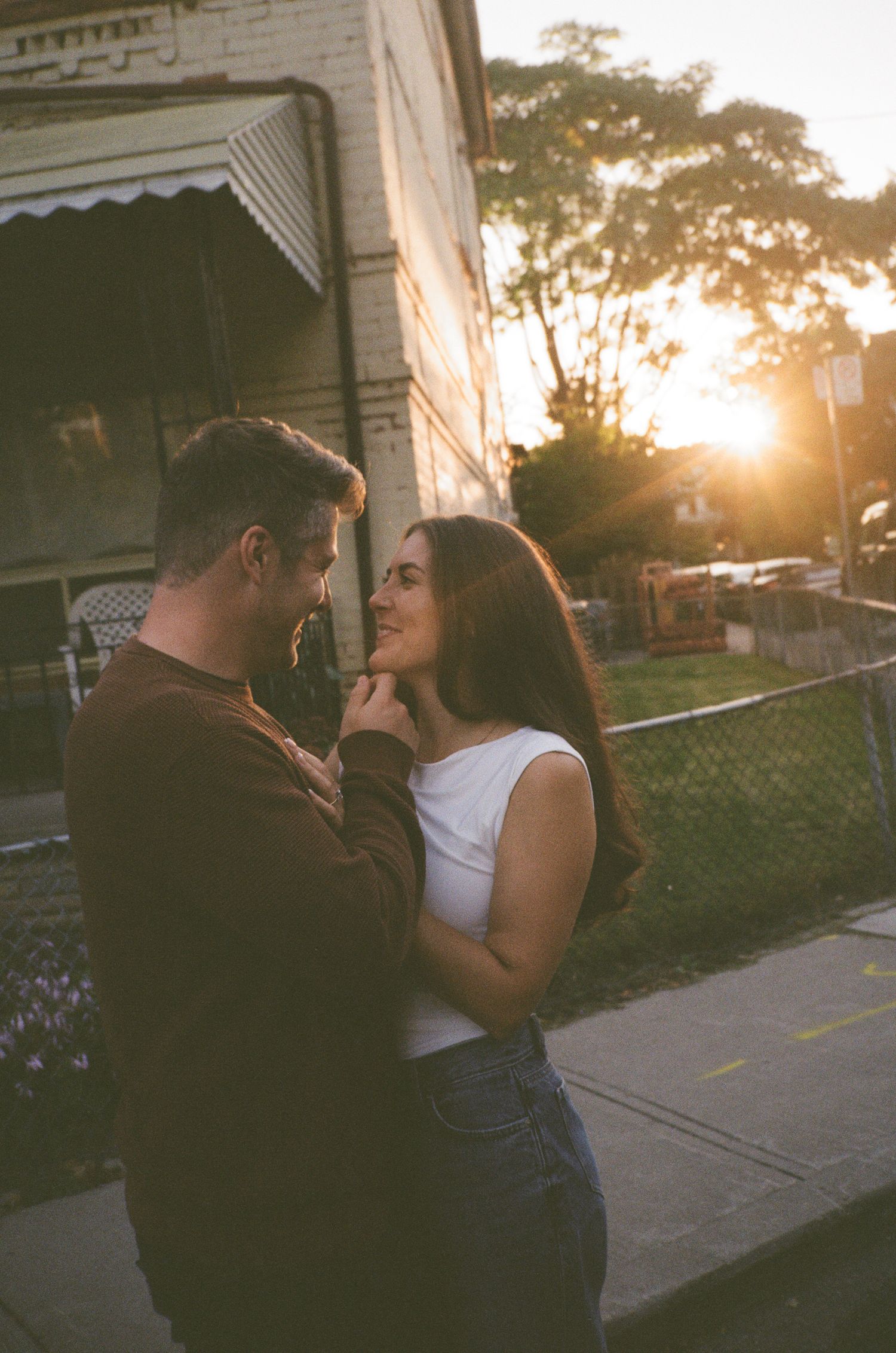 A couple shares an intimate moment at sunset on a sidewalk outside a house with decorative trim.