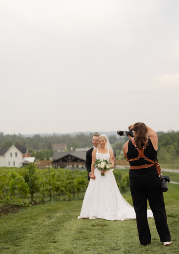 Bridesmaids in black dresses walk through a scenic vineyard with rolling hills in the background on a cloudy day.