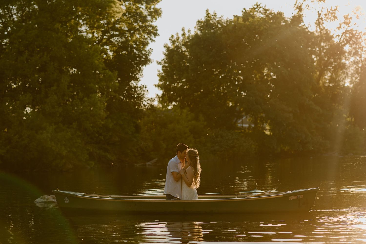 Couple sharing a romantic moment on a canoe at golden hour, surrounded by lush green trees reflecting on calm water.