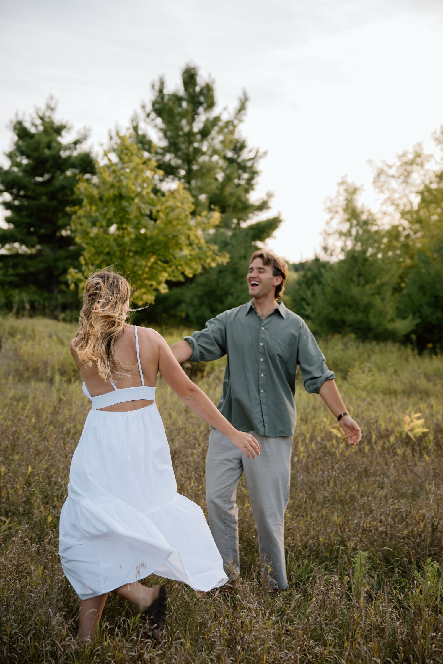 A couple dances together in a grassy meadow at sunset with pine trees in the background.