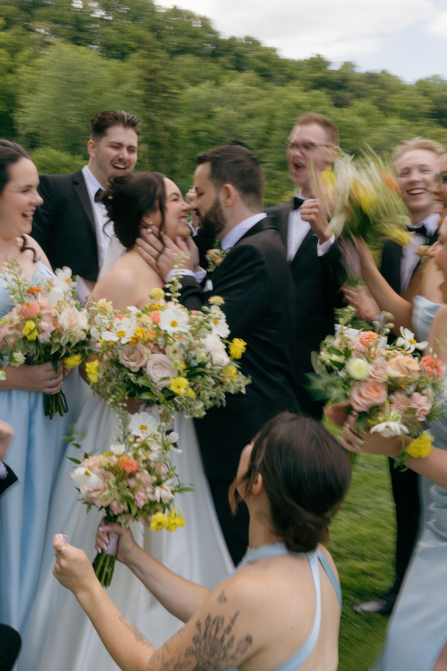 Wedding party in light blue dresses and dark suits laughing together outdoors while holding colorful bouquets.