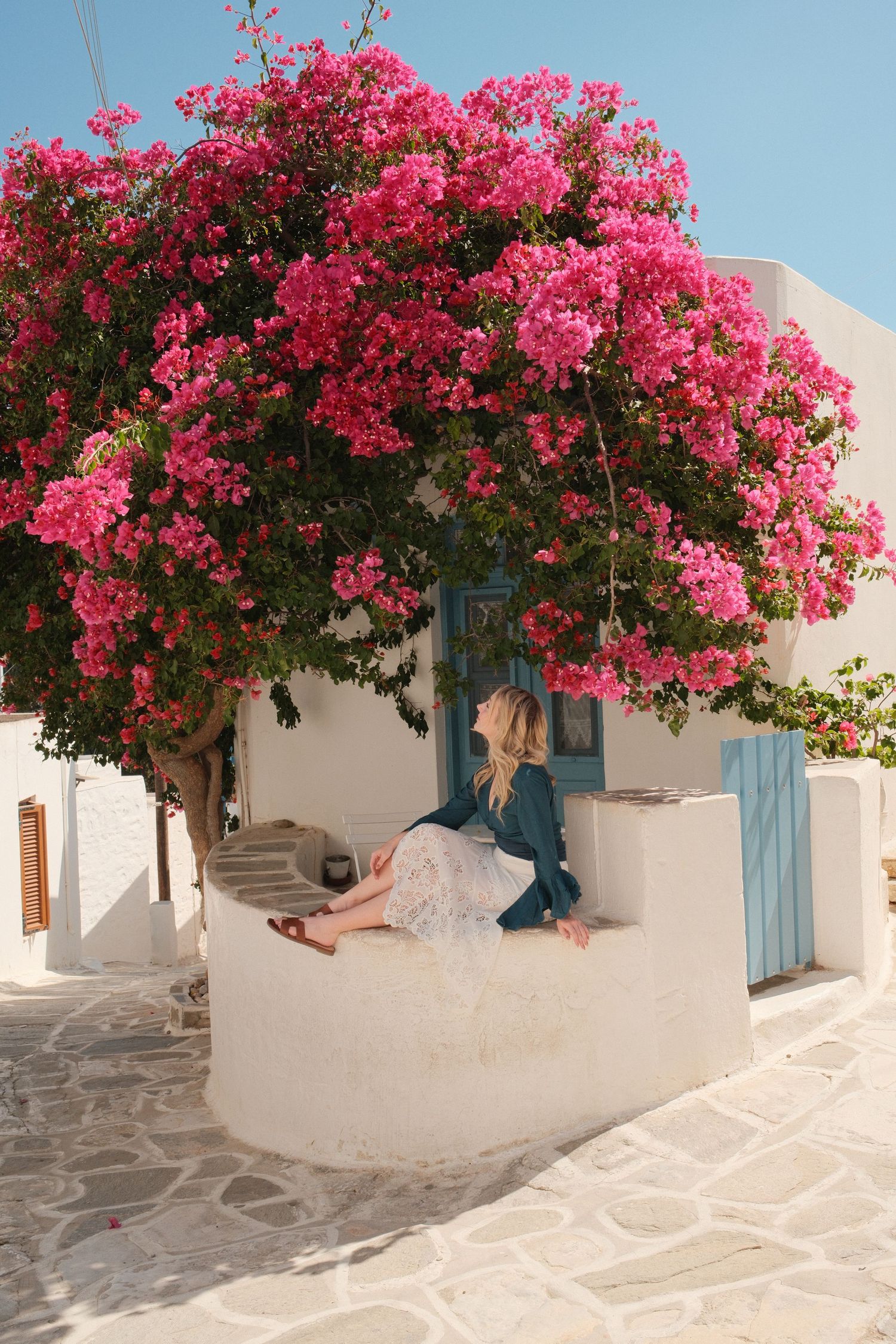Woman sitting under a vibrant pink bougainvillea tree beside a white Greek building with blue door.