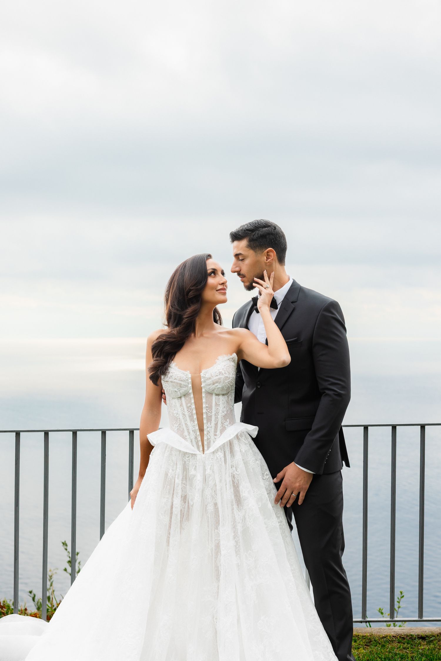 Bride and groom sharing romantic moment in lace gown and black tuxedo at coastal wedding venue.
