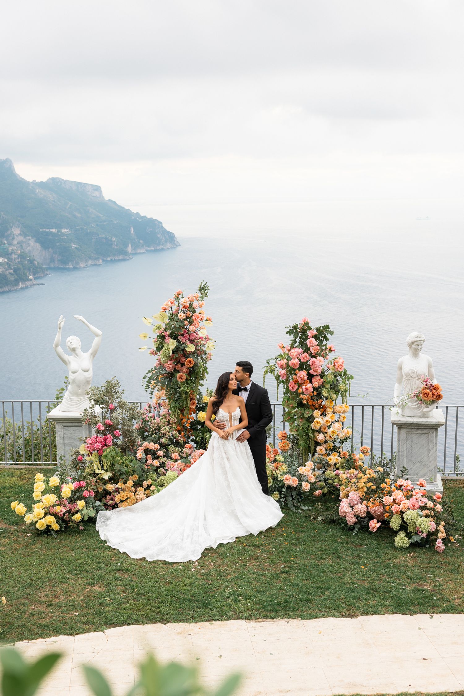 Newlyweds kissing surrounded by vibrant pink, orange, and yellow floral displays with sea backdrop.
