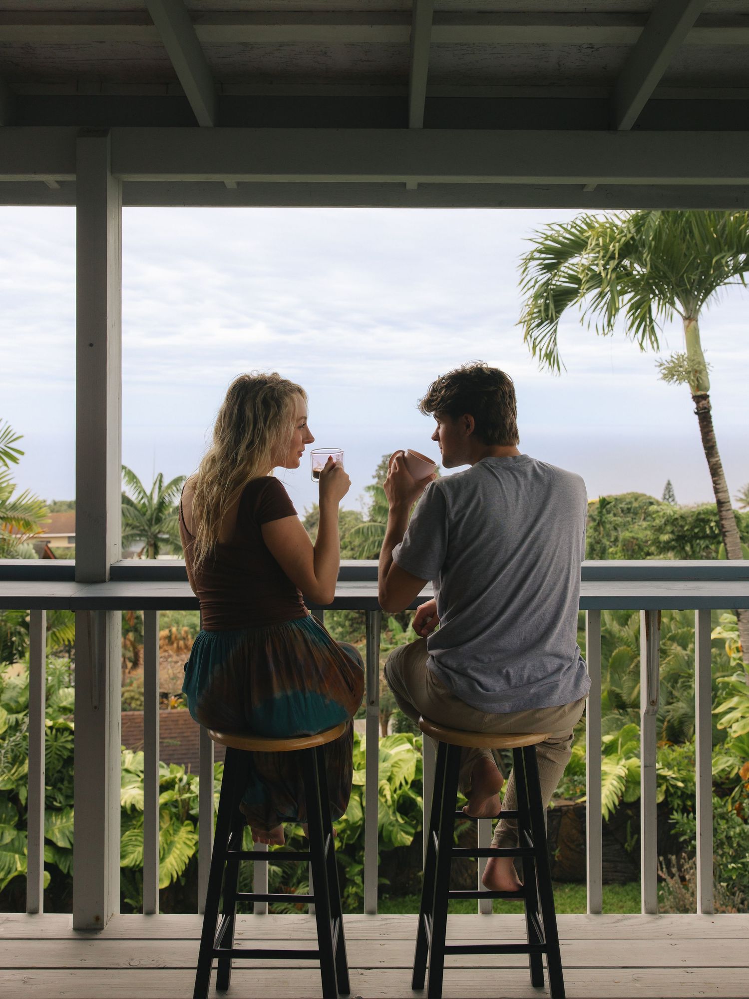 Couple sitting on barstools on a tropical porch, sharing drinks with ocean and palm tree views.