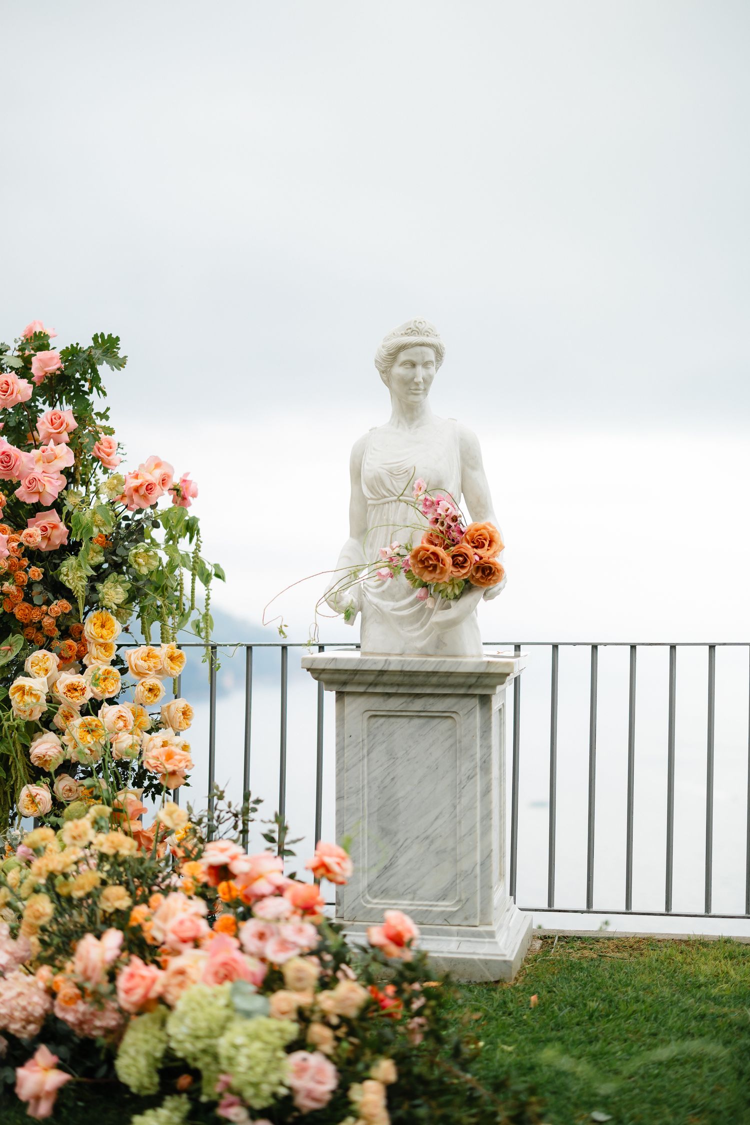White marble statue holding orange bouquet surrounded by colorful peach and pink roses at wedding.