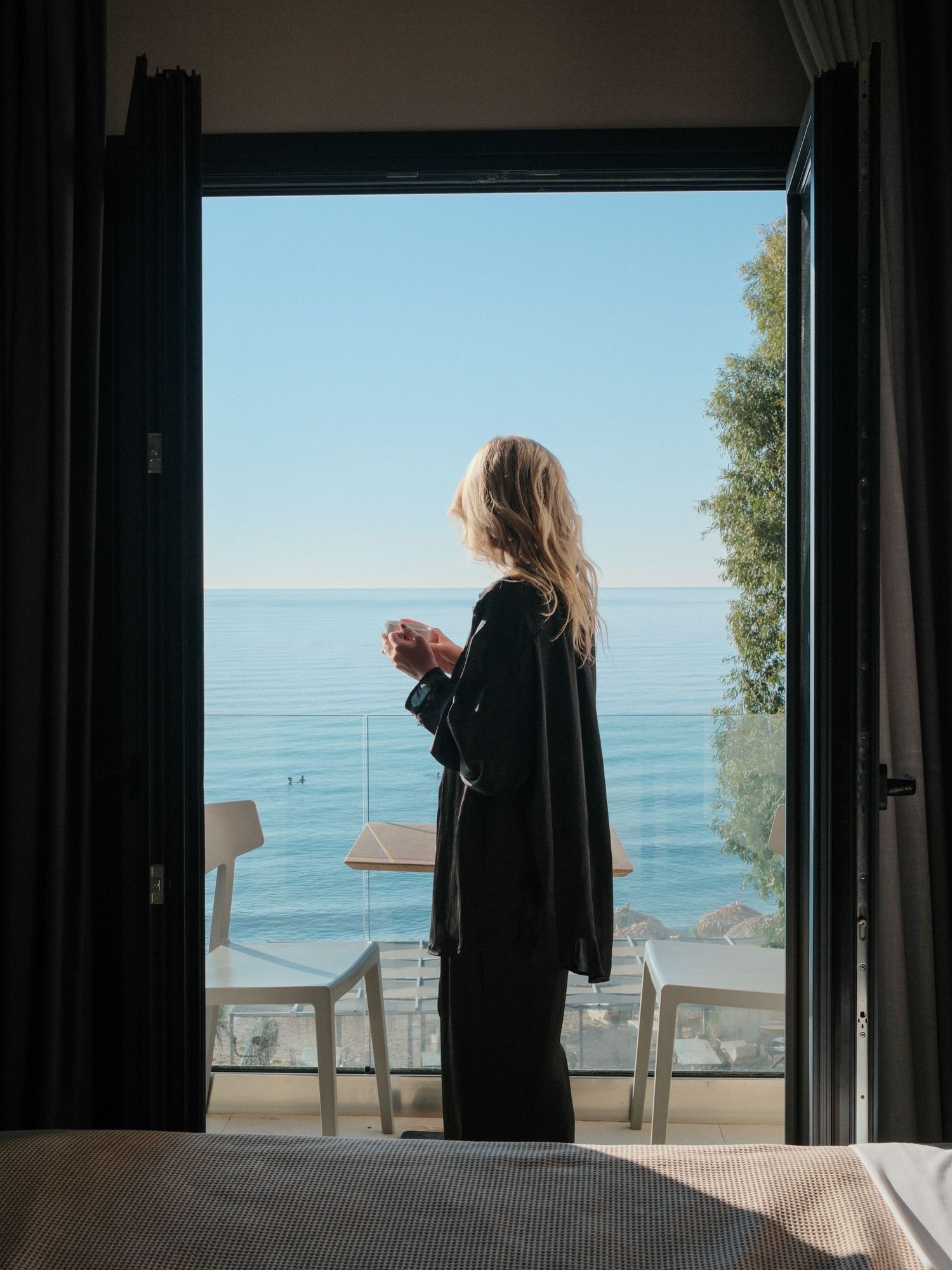 Woman in black outfit stands at open balcony door holding coffee, overlooking a serene blue ocean view.