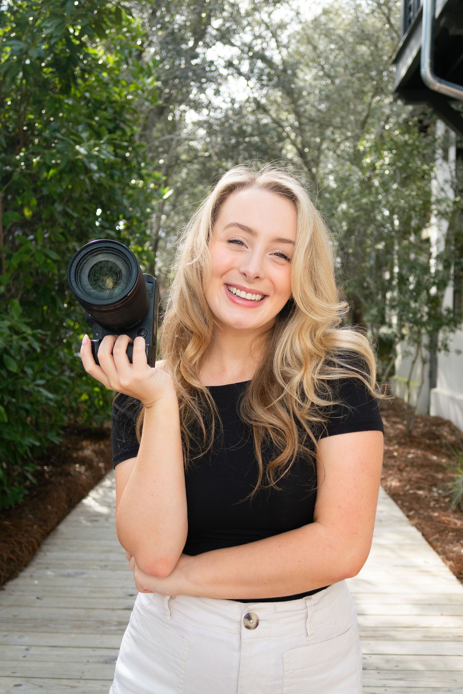 Smiling blonde woman holding a DSLR camera outdoors on a wooden pathway surrounded by lush green trees.