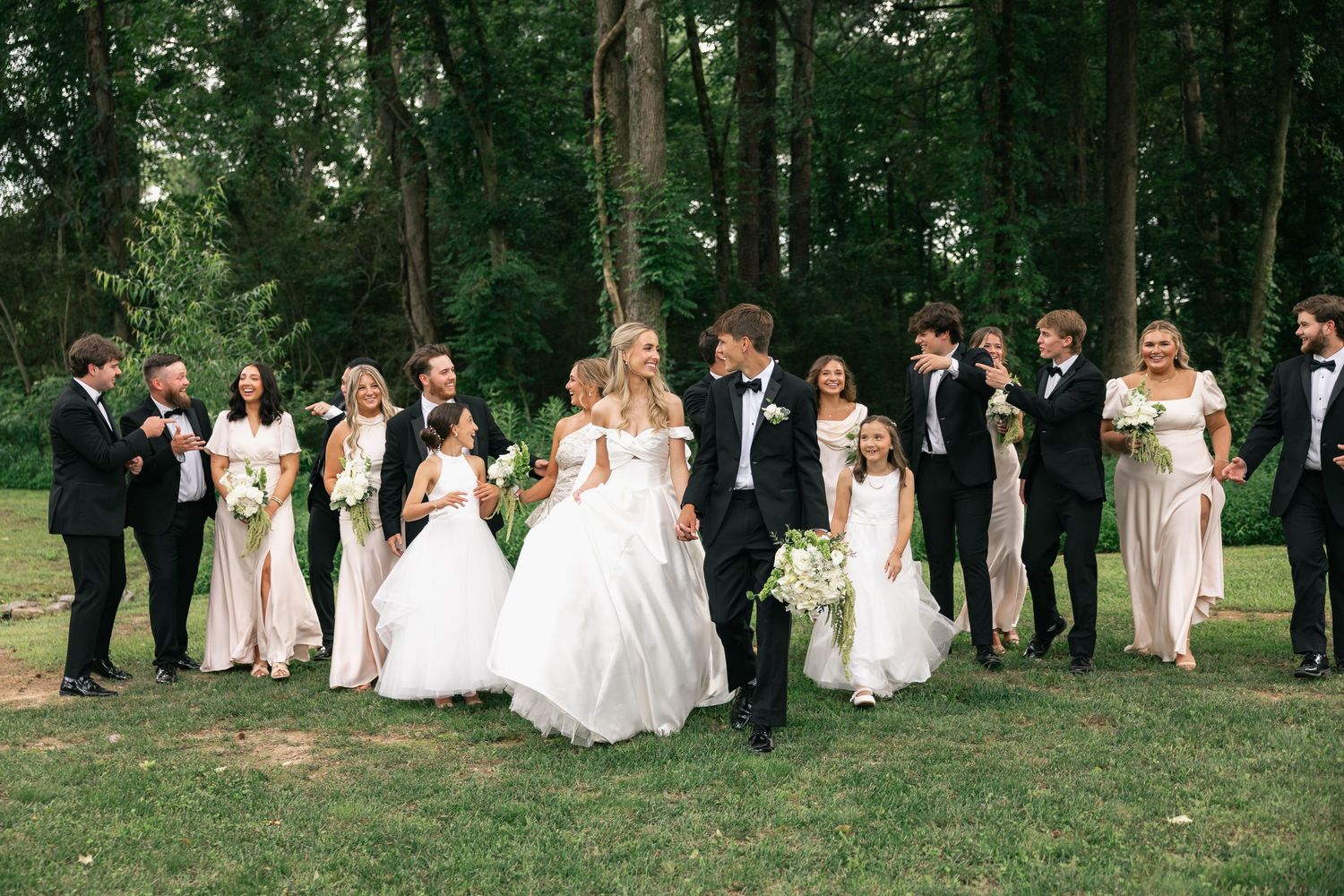 Wedding party poses outdoors with bride and groom kissing, bridesmaids in blush gowns and groomsmen in black tuxedos.