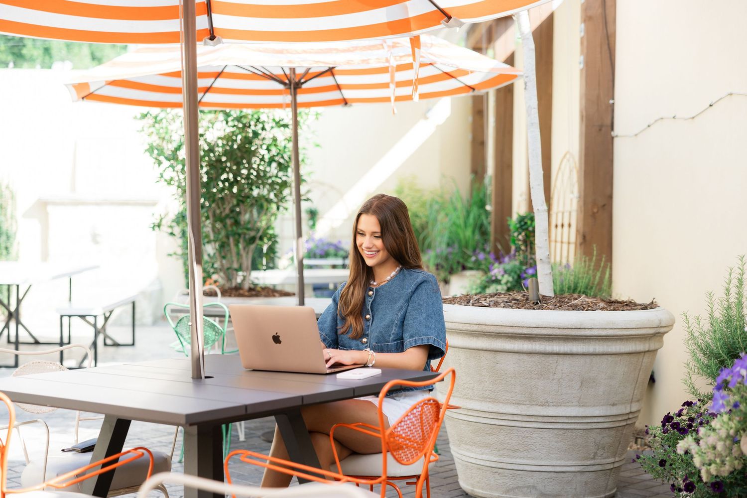 Woman smiling while working on laptop at outdoor cafe patio table under orange striped umbrella.