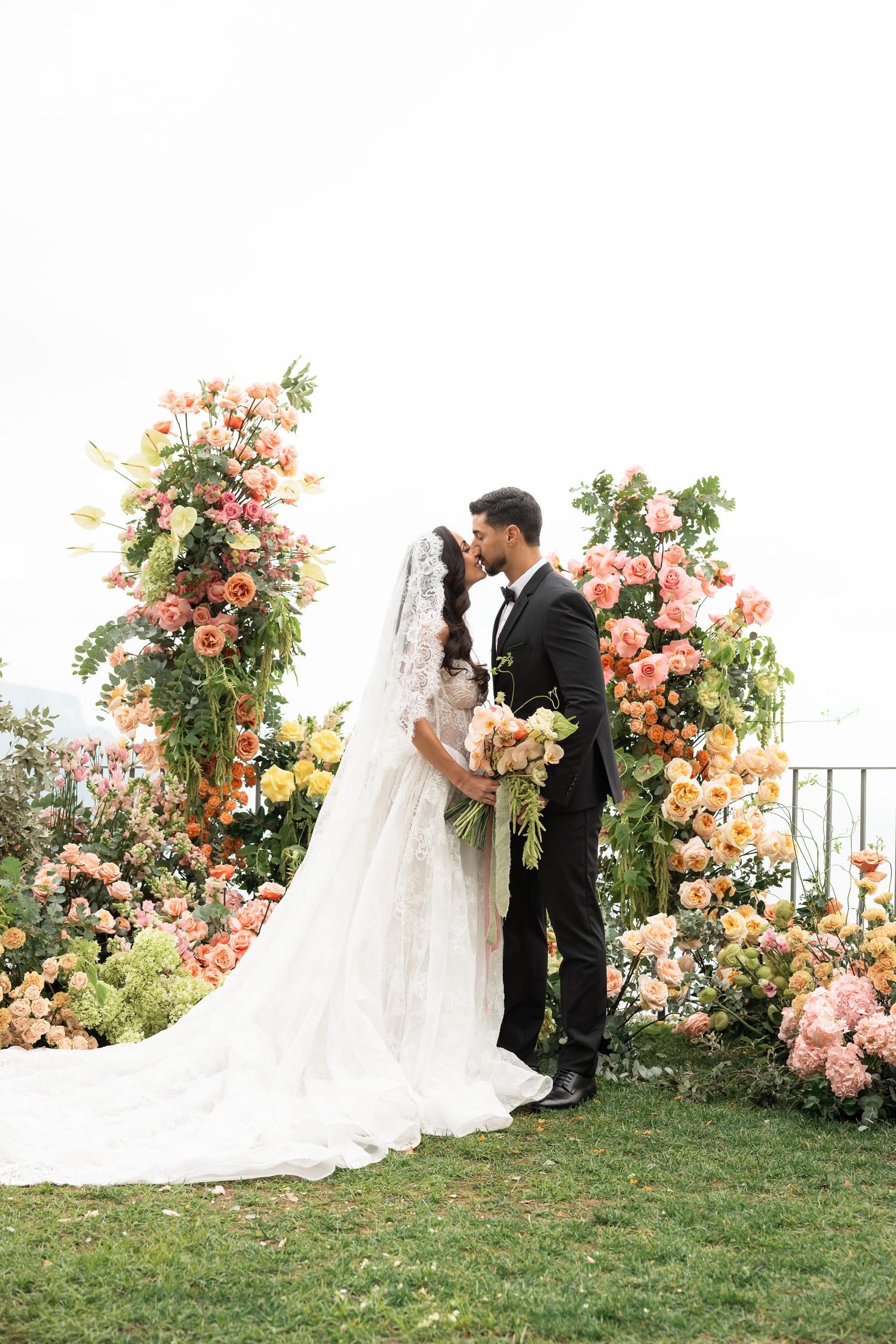 Bride and groom kissing at floral ceremony arch with cascading colorful roses and long lace veil.