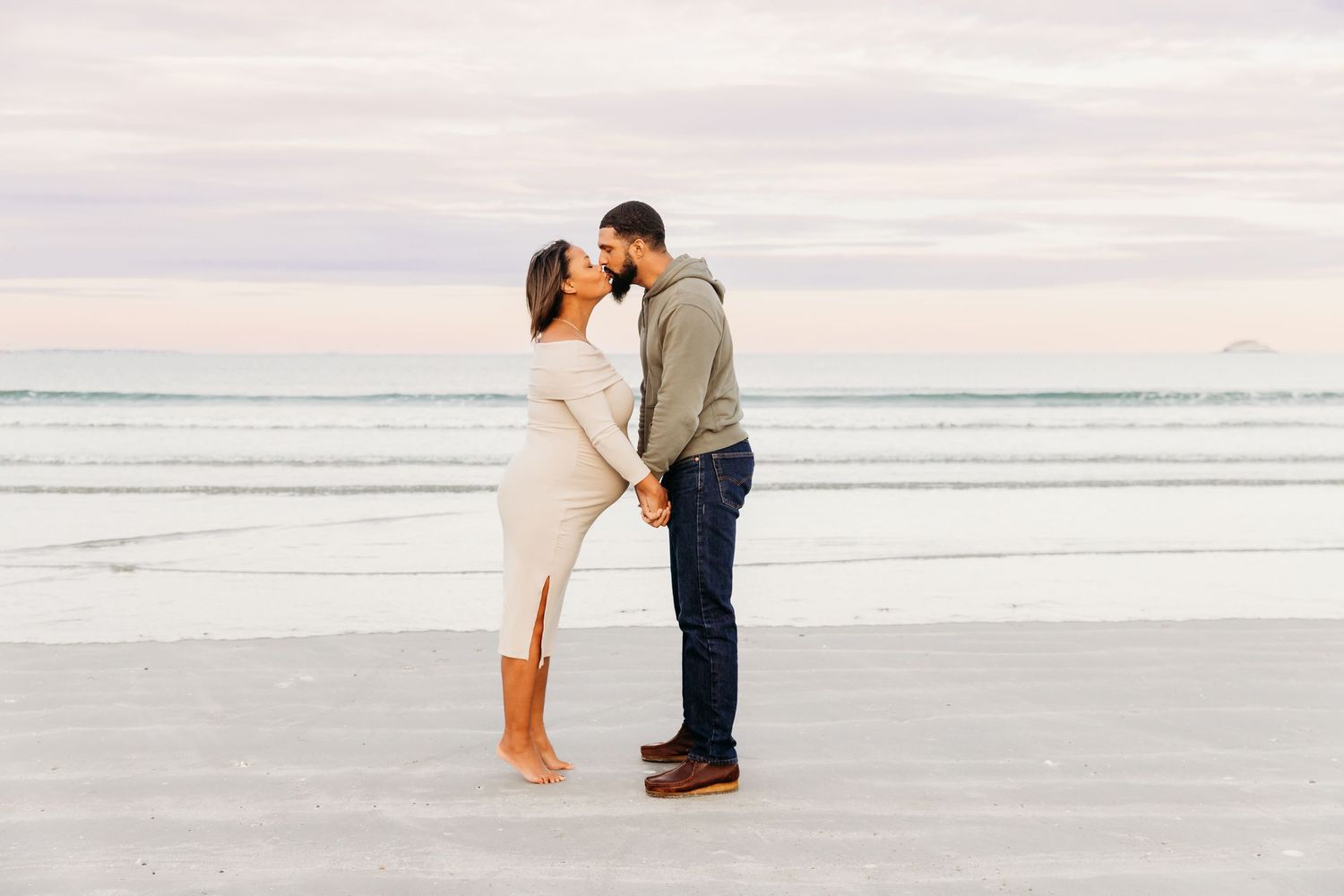 A couple shares a romantic moment on a serene beach at sunset with soft pink skies and ocean waves in the background.