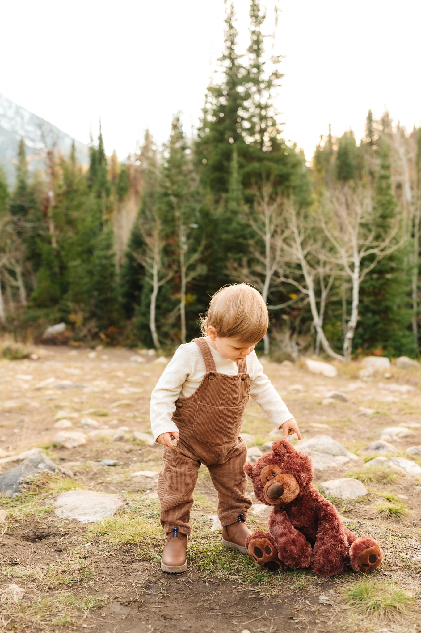 Small child in brown overalls playing with teddy bear on rocky mountain terrain.