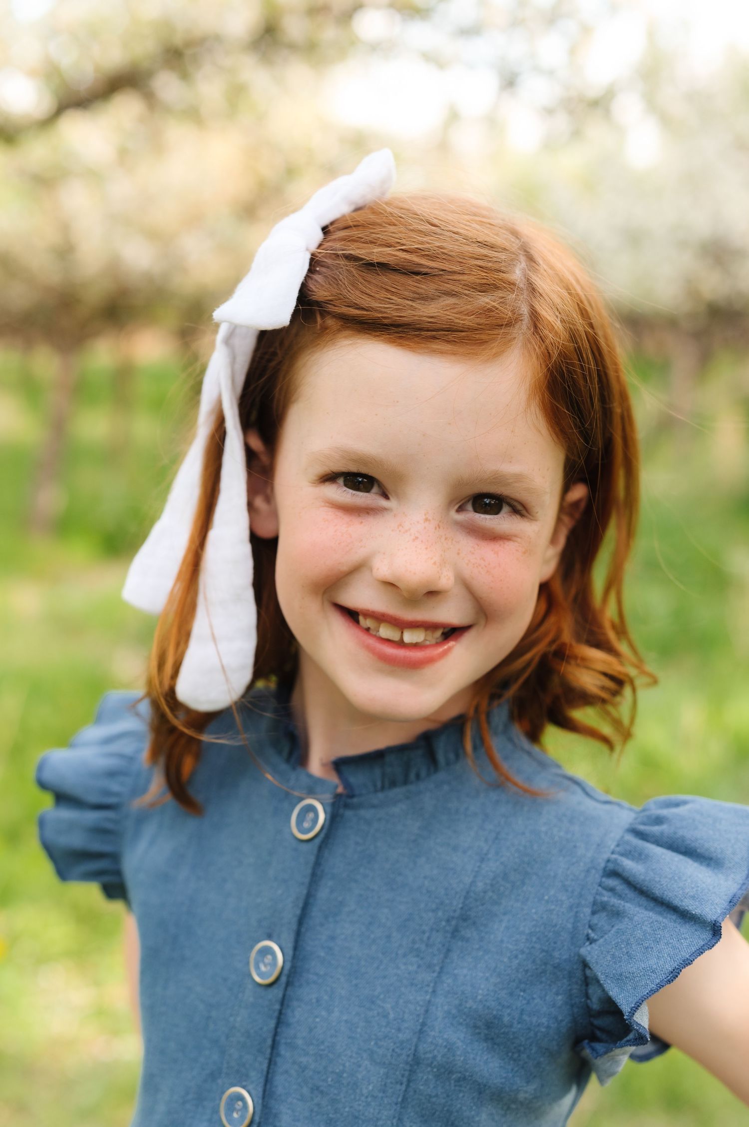 A child wearing a denim dress and white hair bow smiles brightly in a blooming spring orchard.