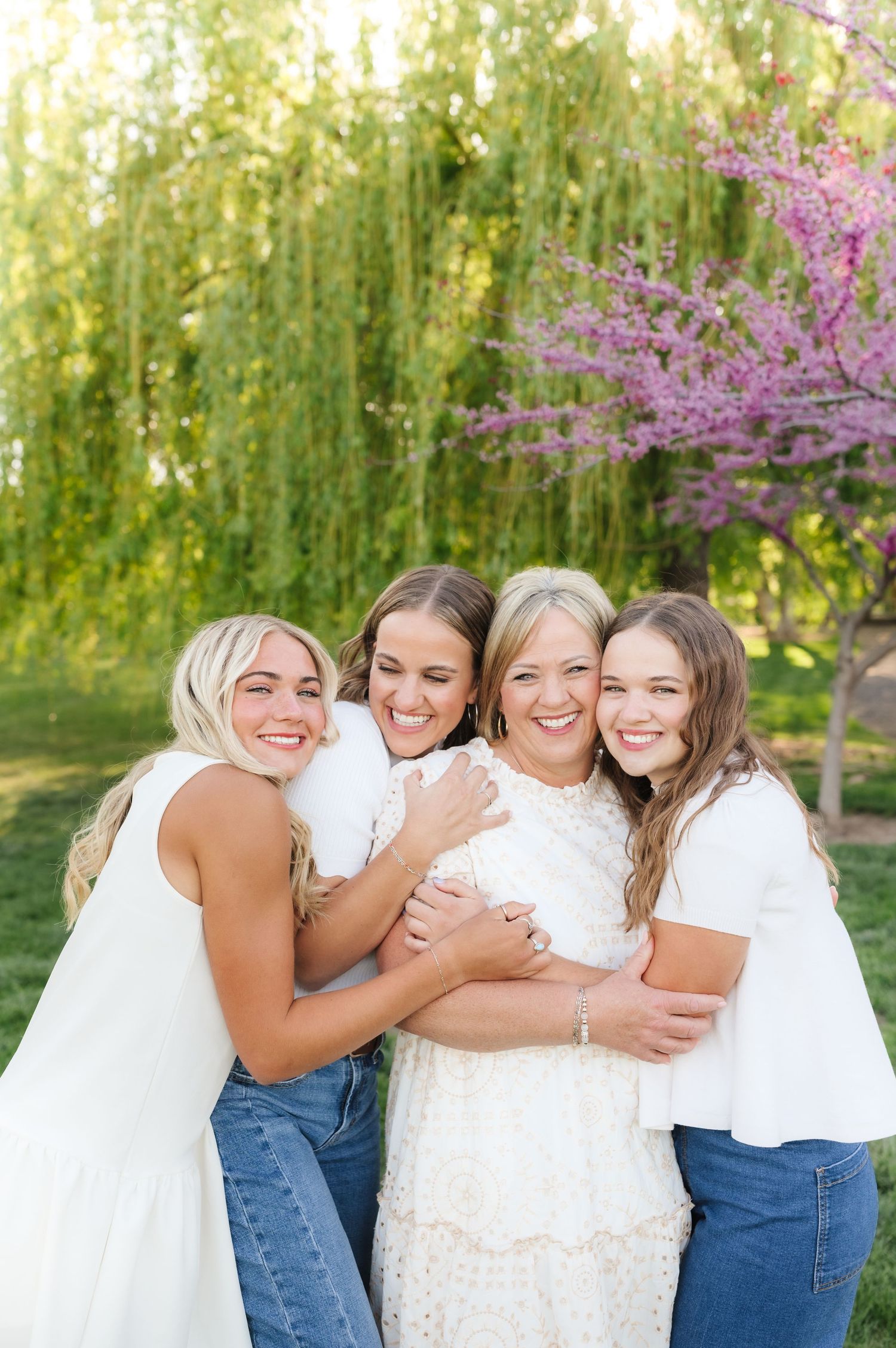 A group in white dresses and denim embrace joyfully in a spring garden with blooming pink cherry blossoms.