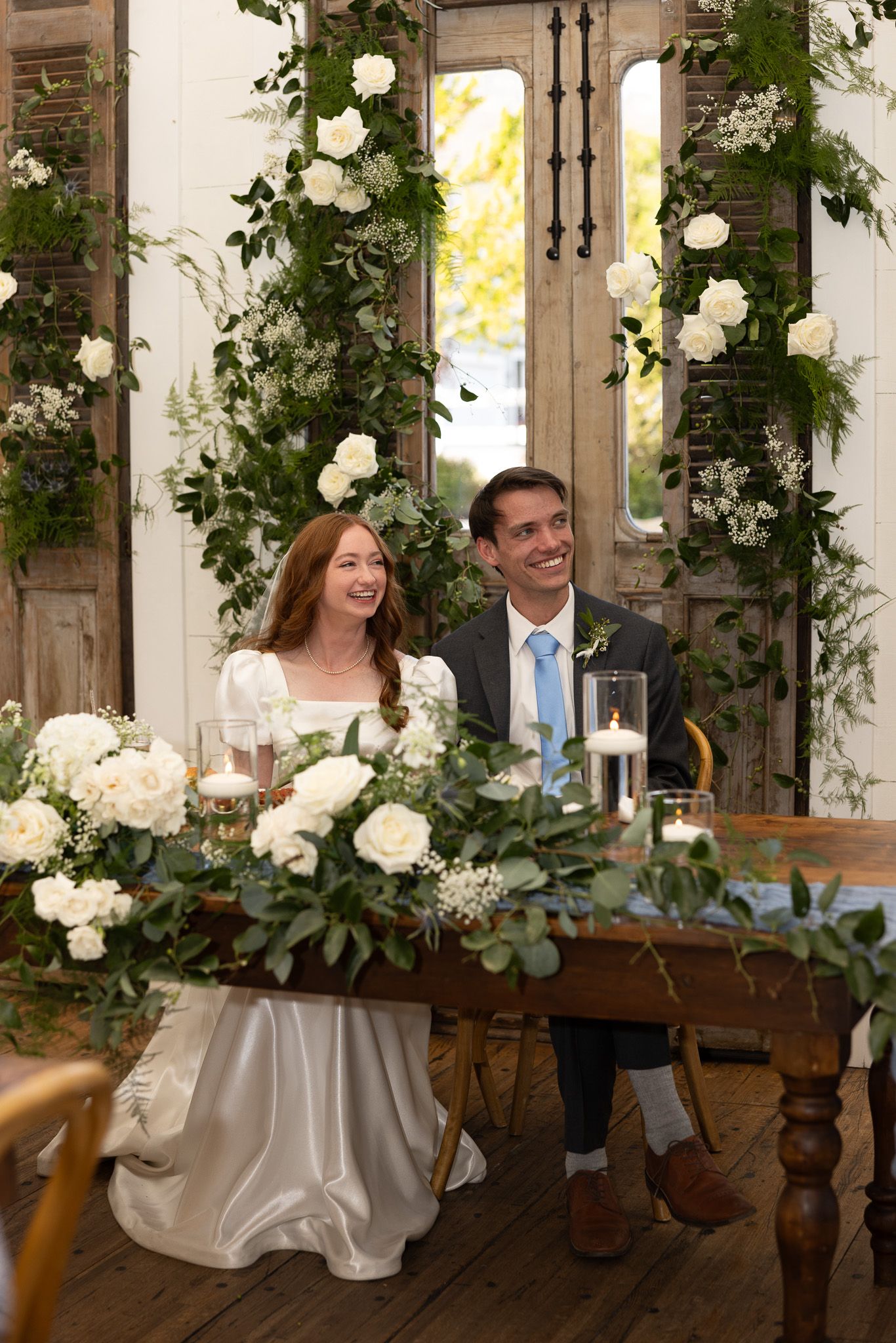 Bride and groom smiling at sweetheart table adorned with white roses and greenery, elegant floral arch backdrop.