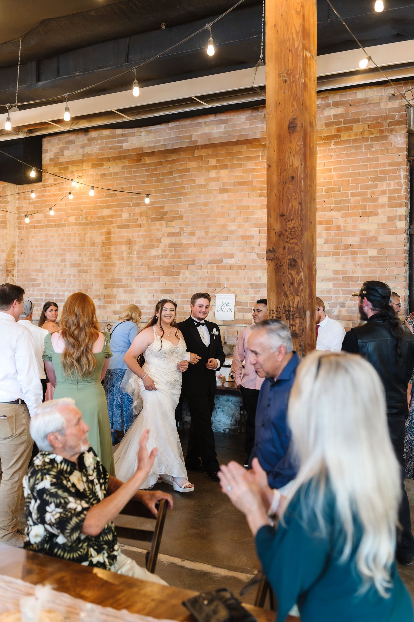 Bride and groom make grand entrance at rustic wedding reception with brick walls and string lights.
