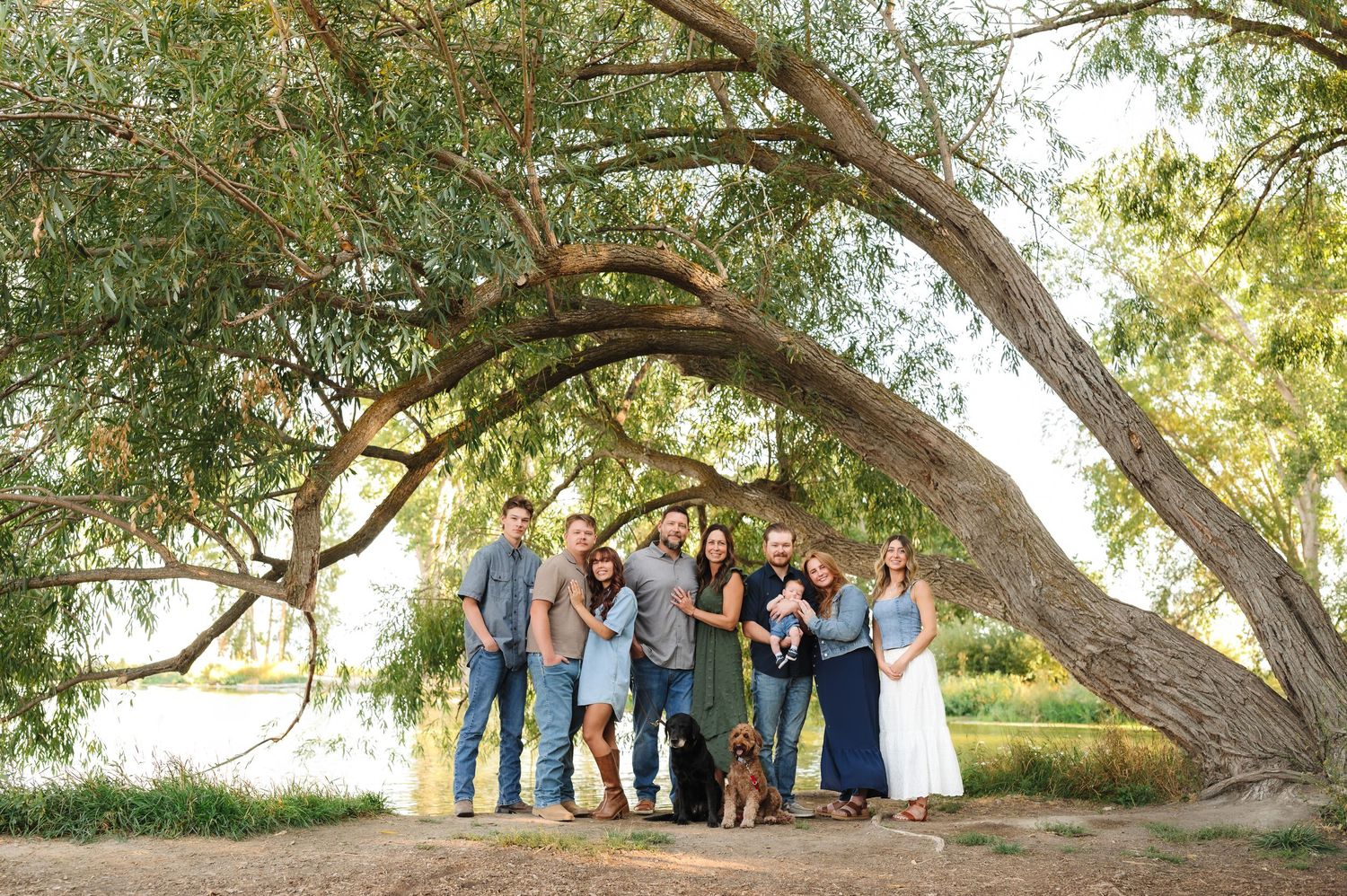 Family portrait of multigenerational group with dog under large willow tree near lake on sunny day.