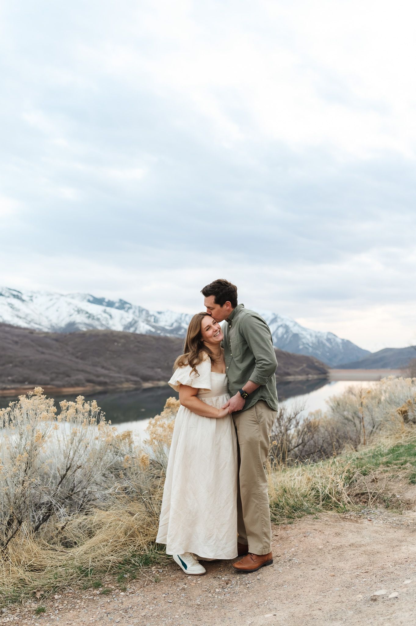 Couple embracing on a scenic overlook with snow-capped mountains and a lake in the background.