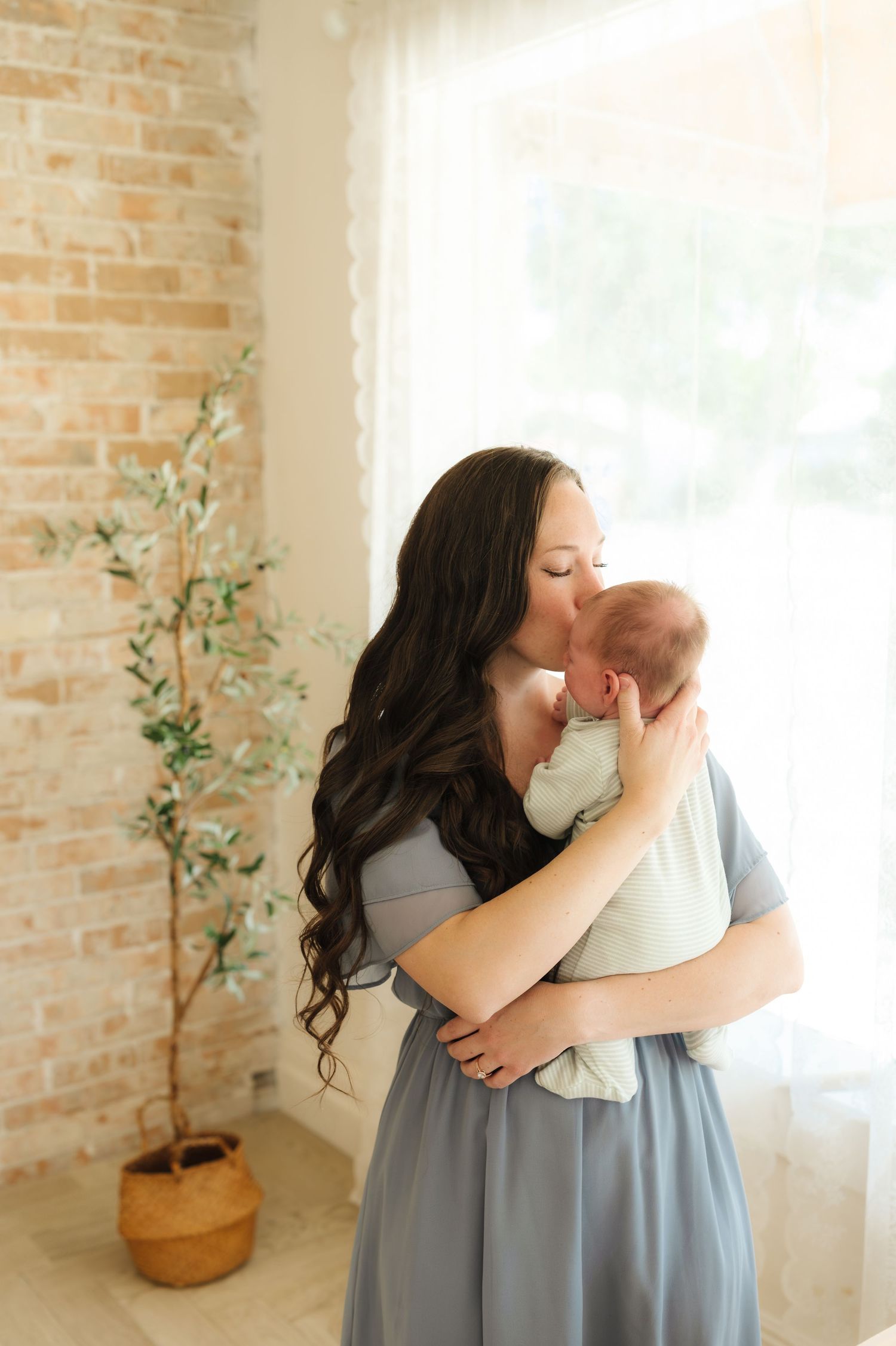 Mother in blue dress embracing baby in white against brick wall with indoor plant.