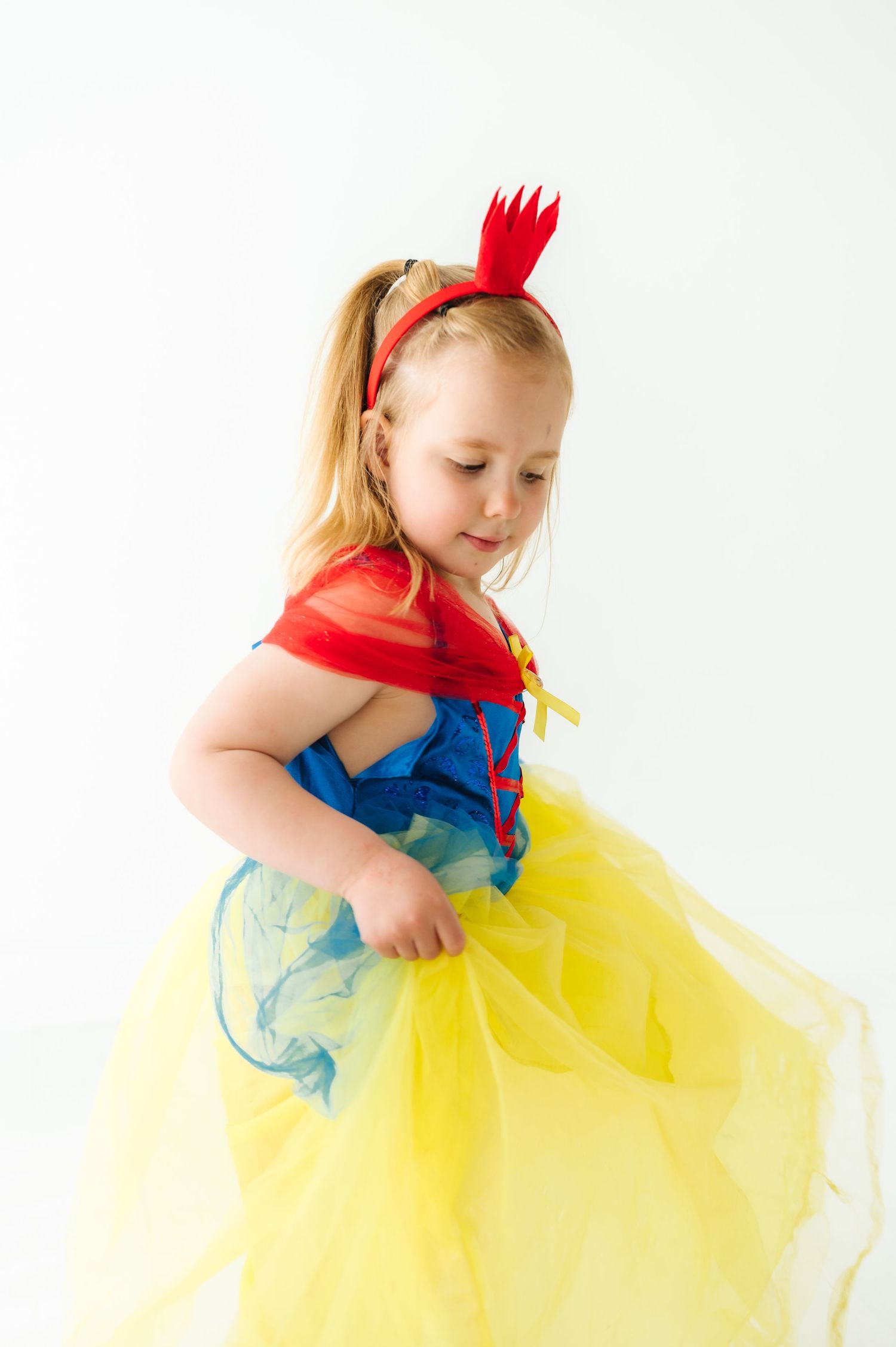 Child twirling in a yellow and blue princess dress with red bow accessory.