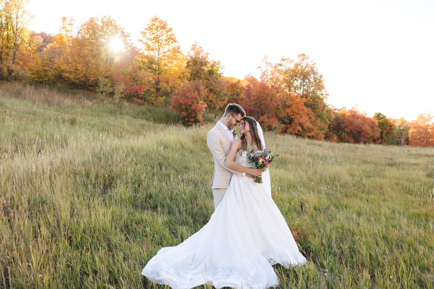Bride and groom embrace in a sunlit autumn meadow, surrounded by colorful fall foliage during golden hour.