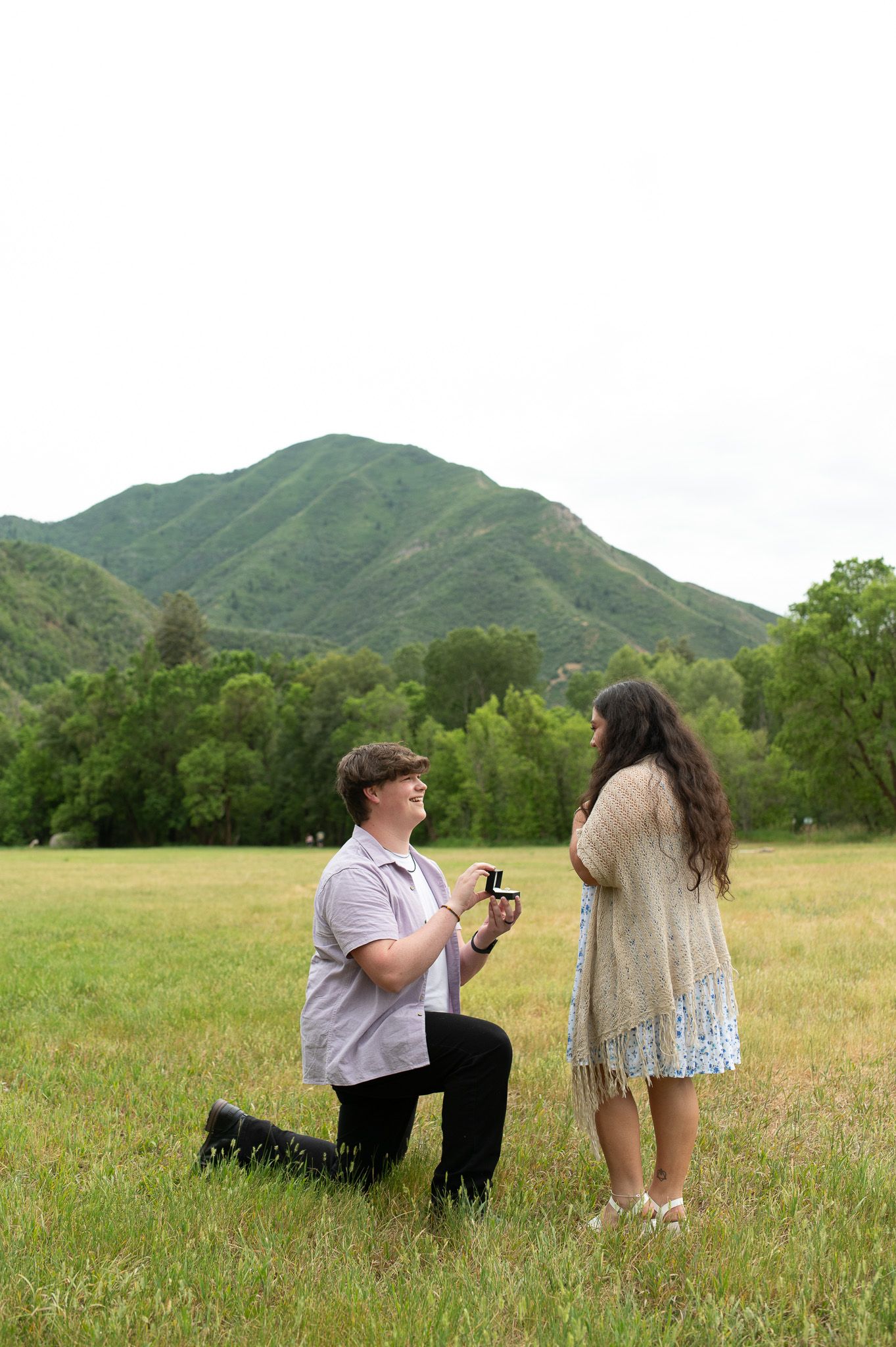 Man kneeling on one knee proposing to woman in green mountain meadow with ring box.