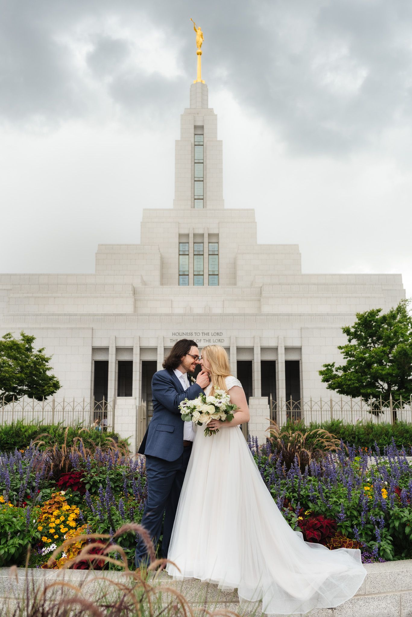 A romantic wedding couple poses in front of the white marble Idaho Falls Temple with colorful flower gardens in bloom.