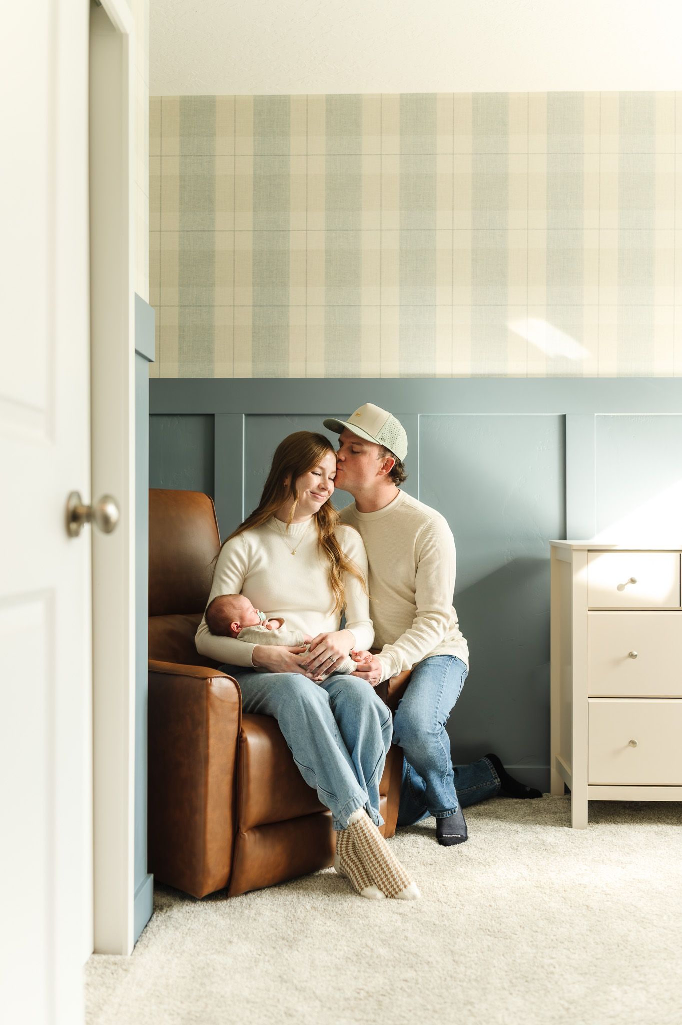 Couple sits together in a nursery chair, smiling and holding their newborn baby in a cozy, softly decorated room.