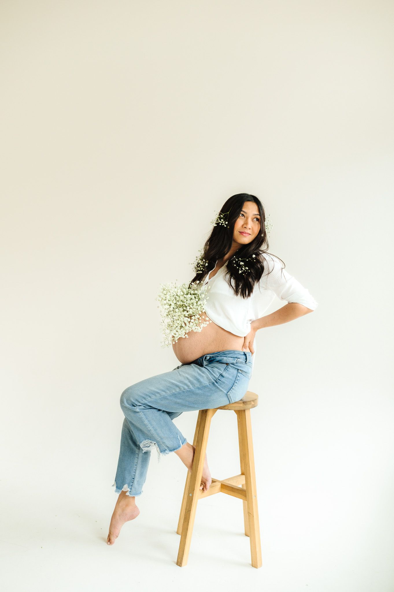 Maternity photo of woman in jeans and white top sitting on wooden stool holding baby's breath flowers.
