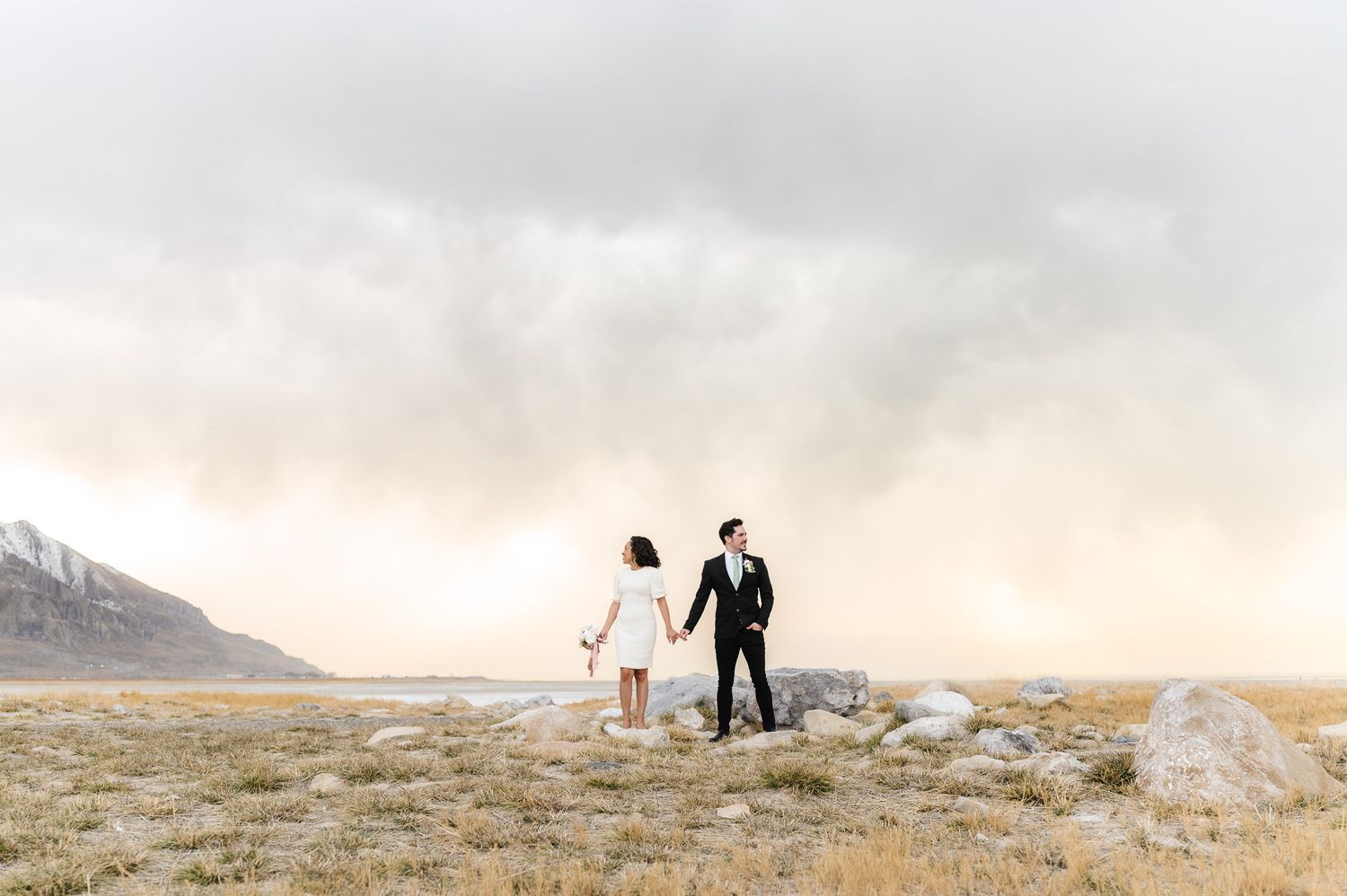 Silhouettes of wedding couple walking across scenic mountain landscape at sunset.