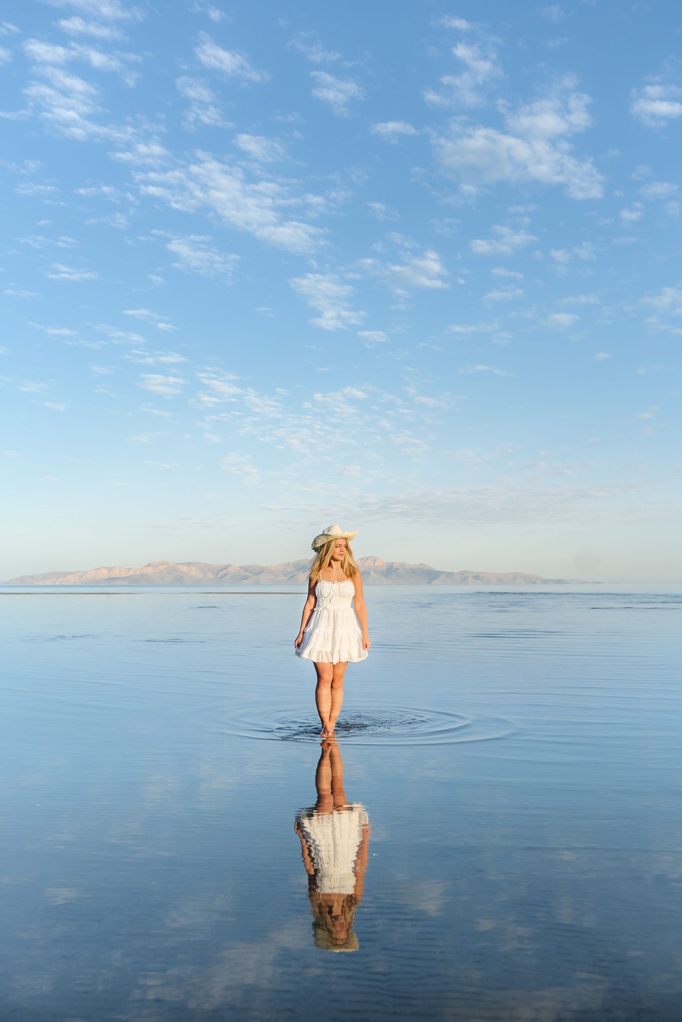 A person in a white dress stands on a reflective surface surrounded by calm waters and blue skies for her senior session