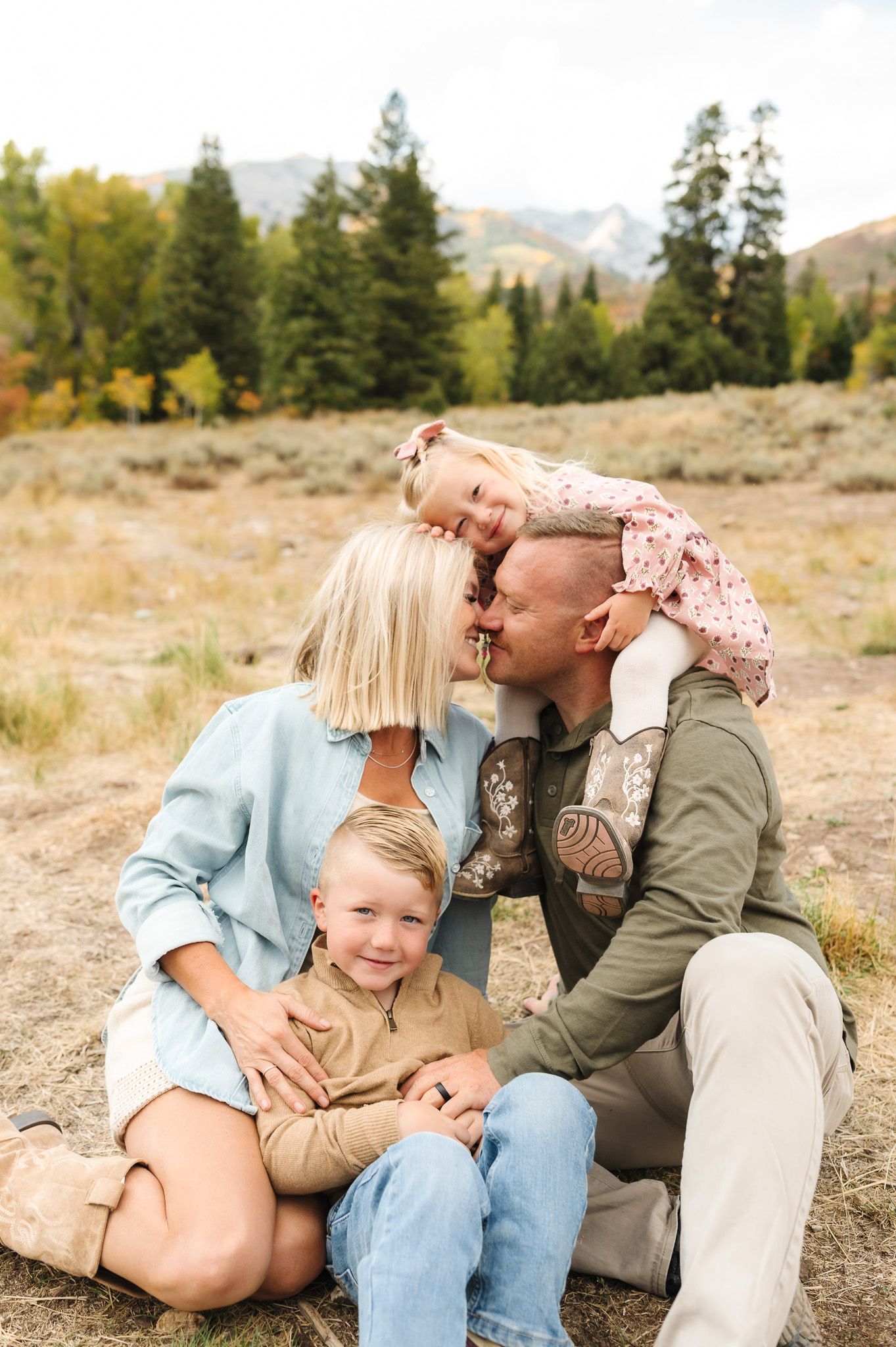 Family shares playful moments together while sitting on ground with mountains and trees in background.