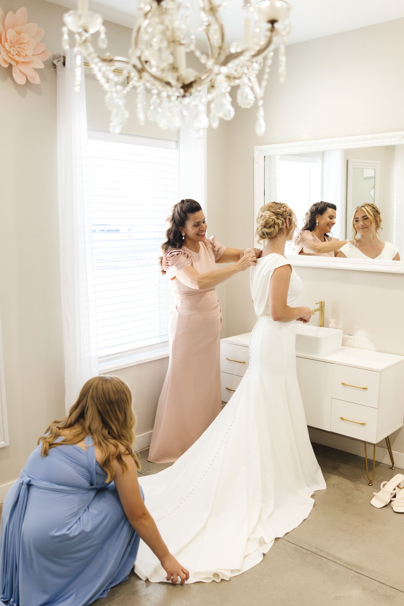 Bridesmaids in blush pink and blue dresses help bride get ready in elegant white dressing room with chandelier.