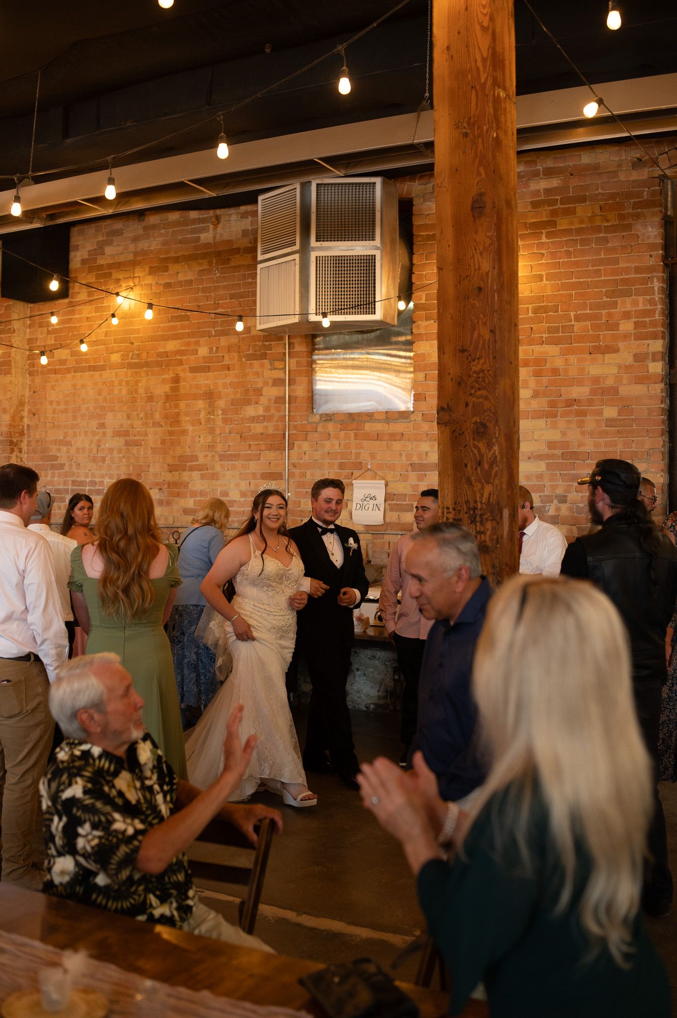 Bride and groom make grand entrance at rustic brick wedding venue with string lights and cheering guests.