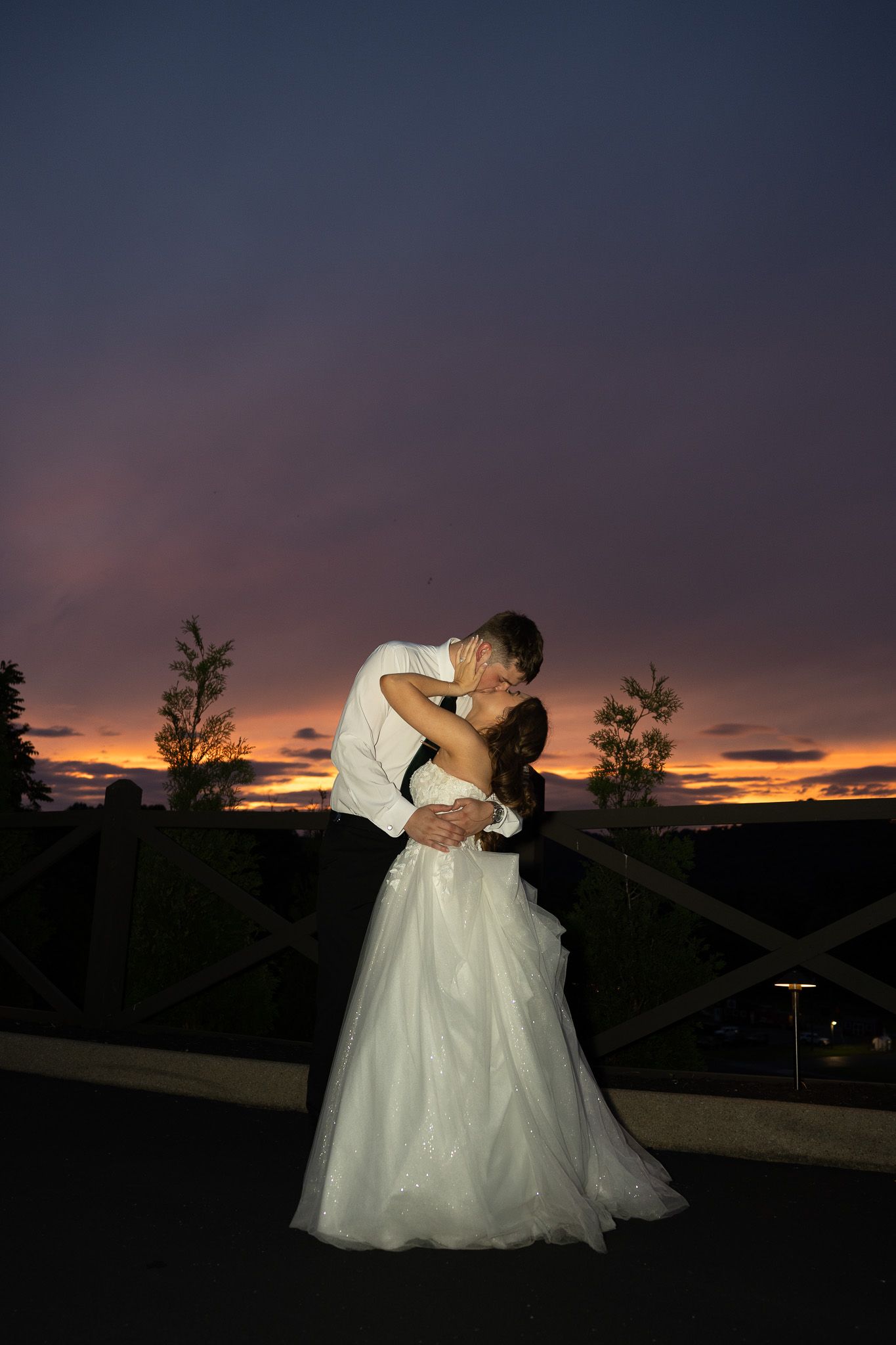 Bride and groom sharing a romantic kiss at sunset, silhouetted against a vibrant purple and orange sky.