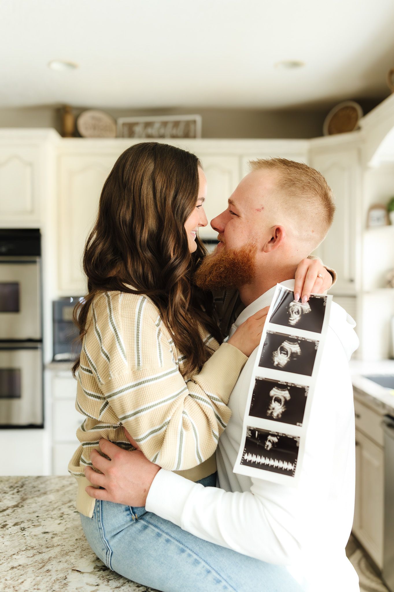 A couple embraces in a kitchen, sharing a tender moment while holding ultrasound photos, announcing their pregnancy.