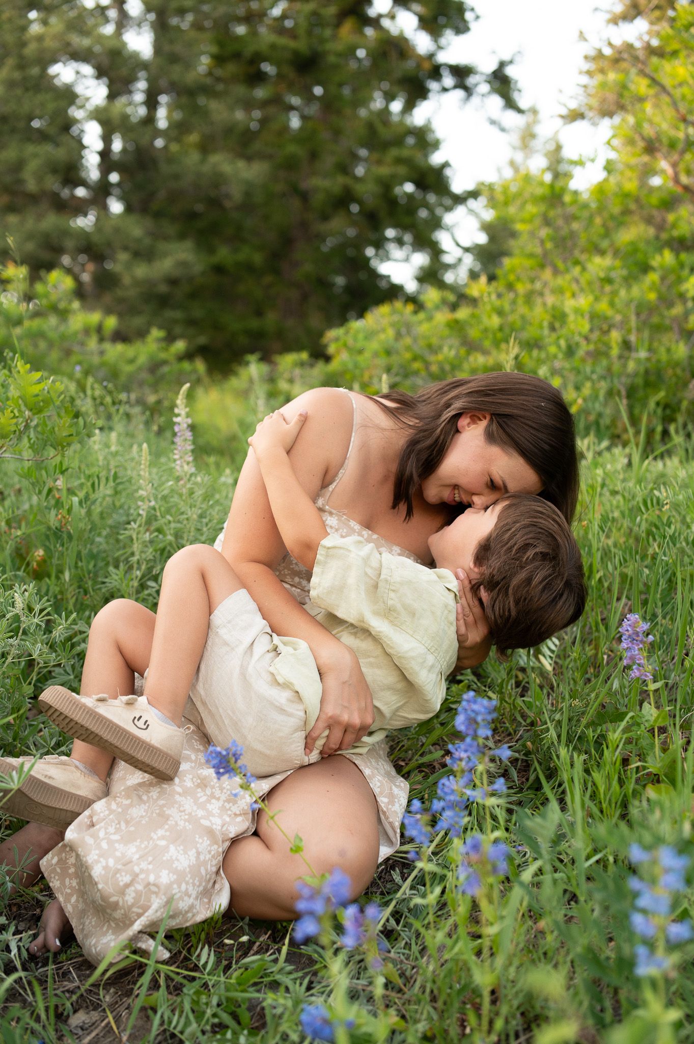 A mother tenderly kisses her young child while sitting in a lush green meadow with wildflowers.