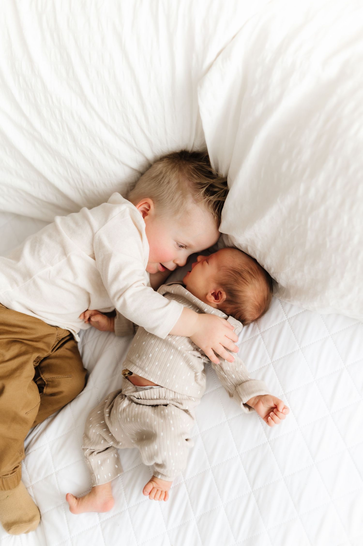 Two siblings in white clothing cuddle together on white bedding during intimate photo session.