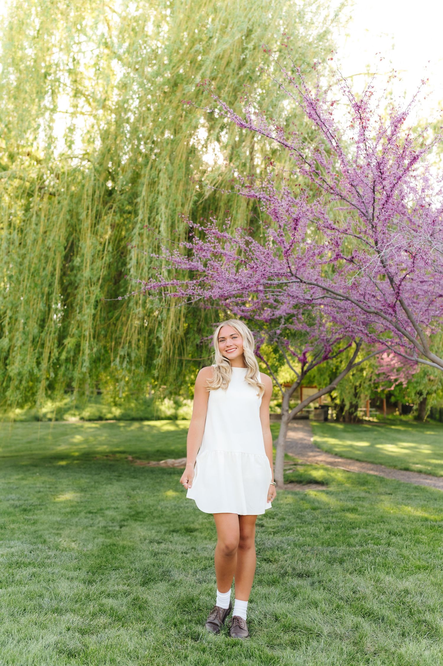 A child in a white dress stands on grass near blooming pink cherry blossoms and weeping willow trees.
