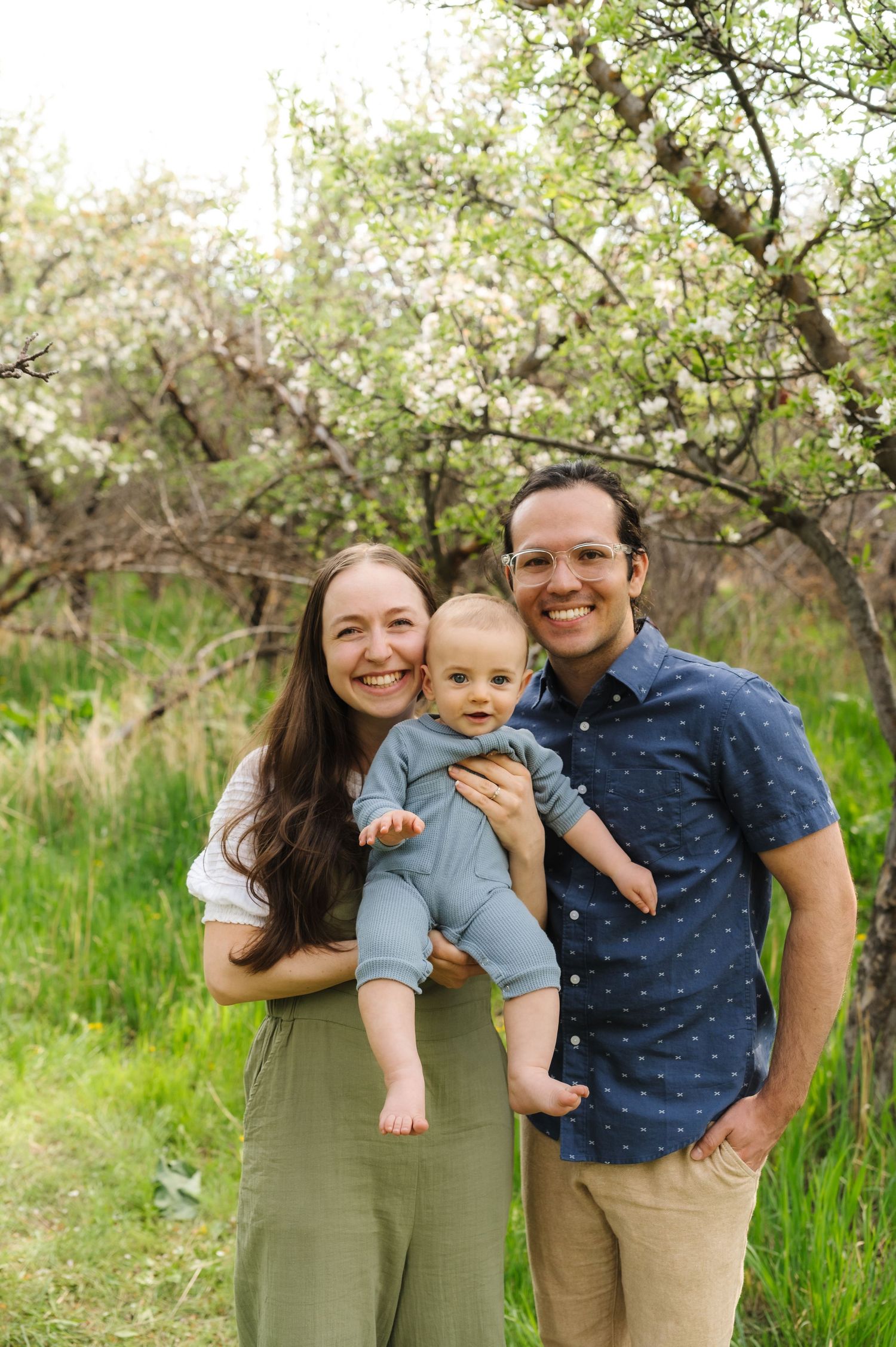 A family of three poses together in a blooming spring orchard with green grass and white blossoms in the background.
