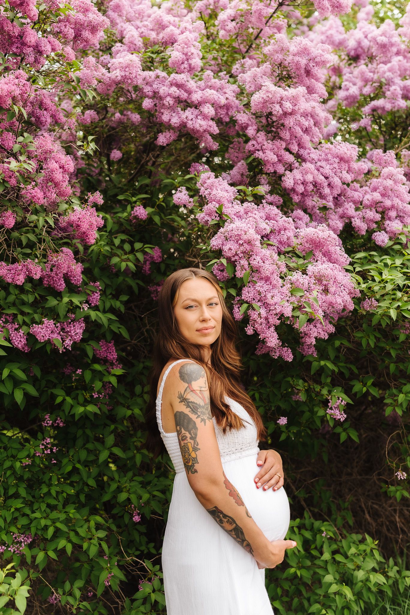 Expectant mother in white dress poses beside vibrant purple lilac blossoms.