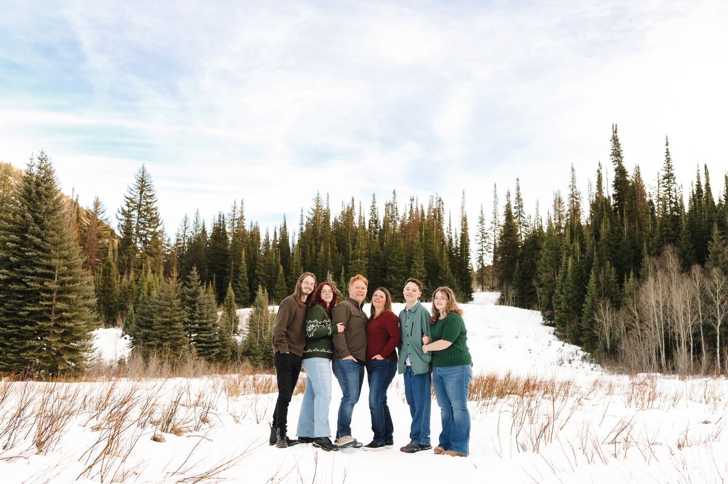 Five women posing together in a snowy winter landscape with evergreen trees under a blue sky.