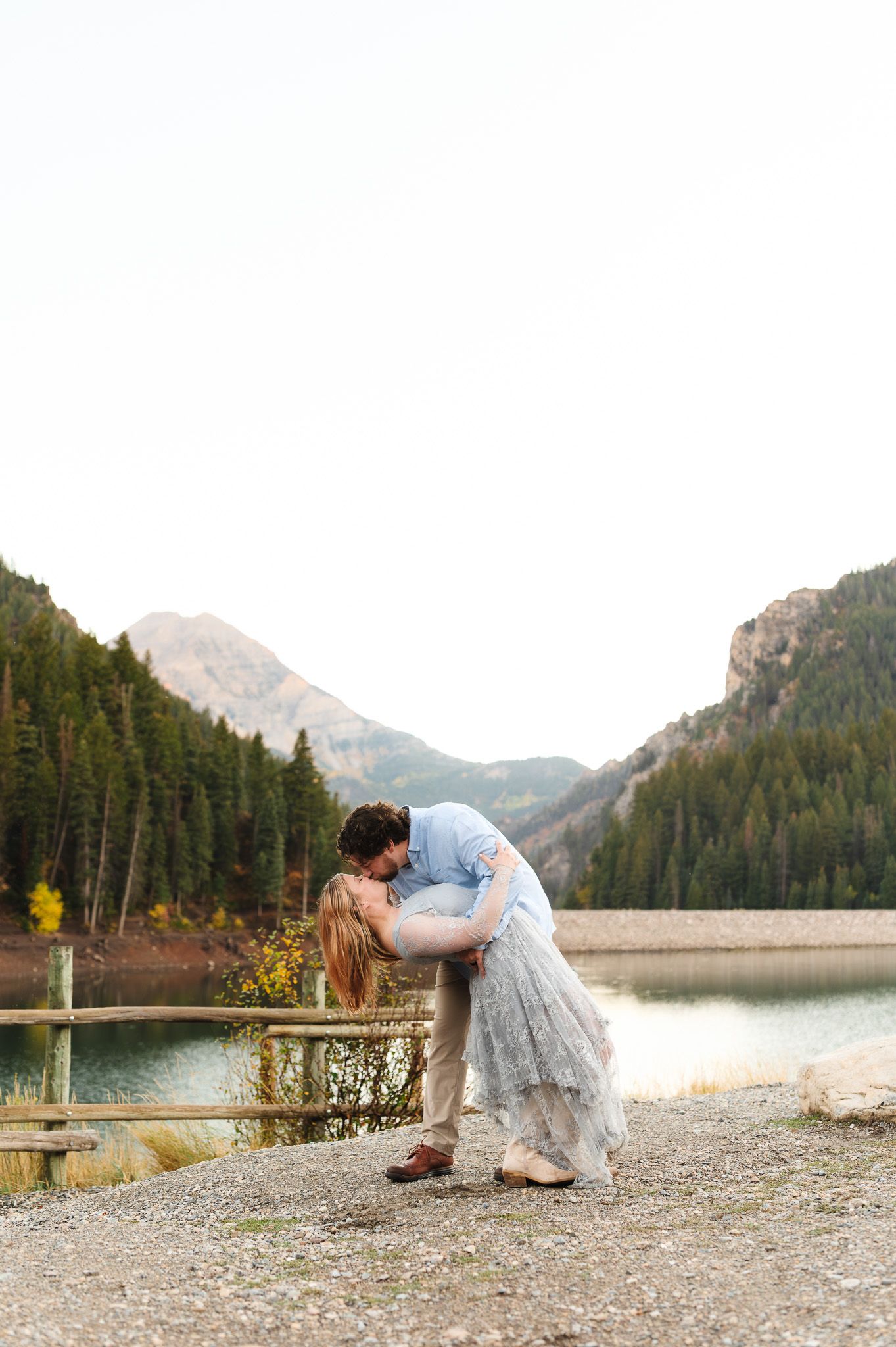 Couple sharing a romantic dip kiss by a mountain lake with pine trees and peaks in the background.