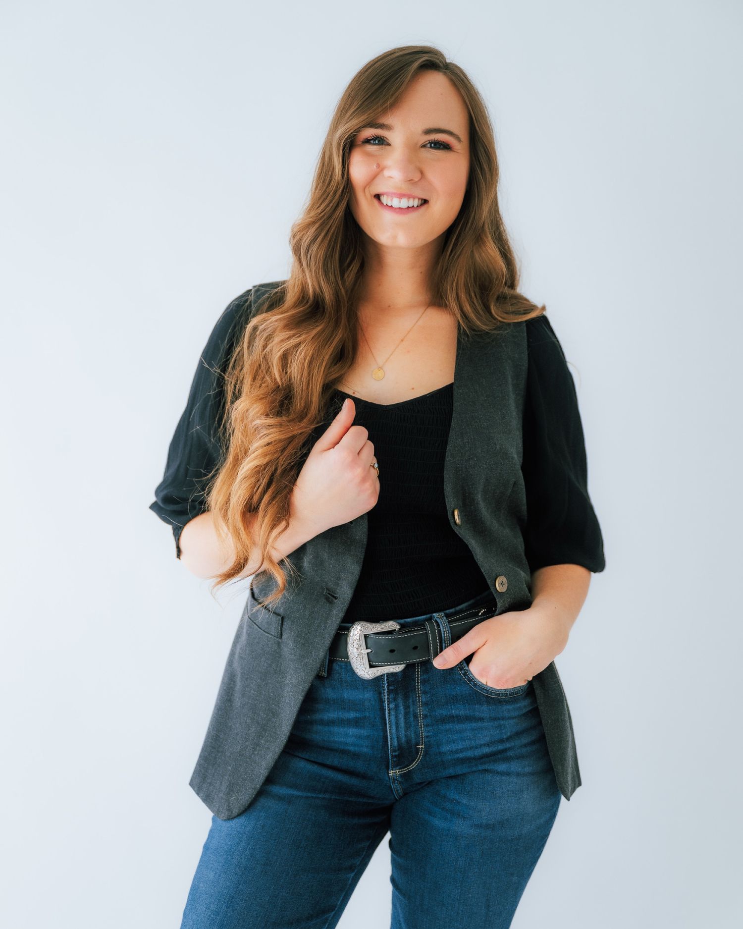 Casual business style outfit featuring dark blazer, black top, jeans and silver belt buckle against white background.
