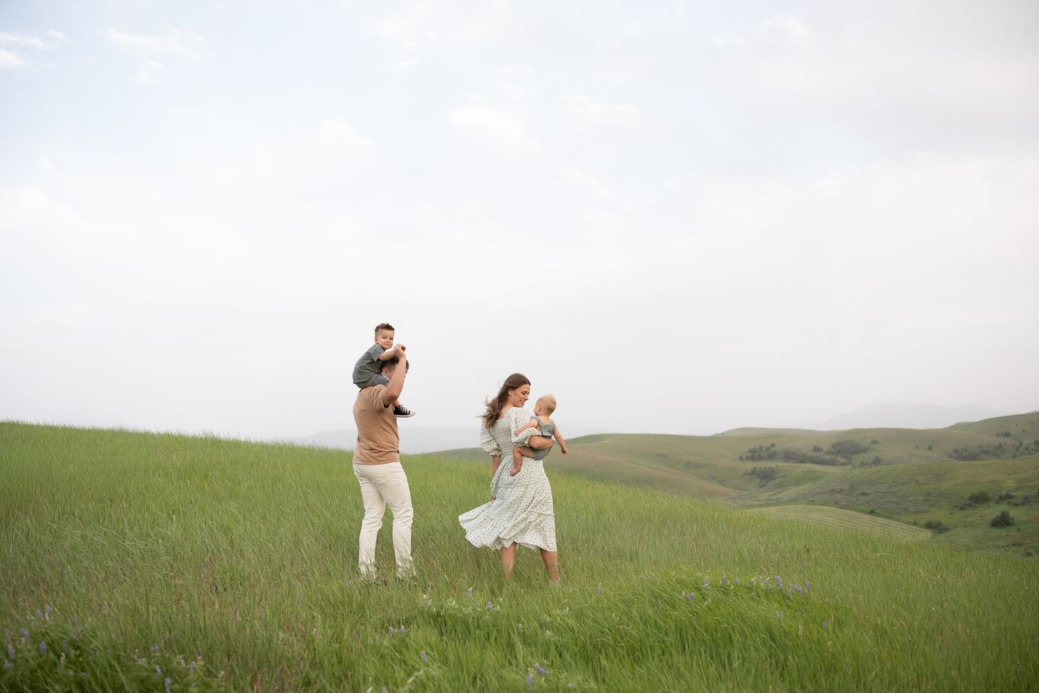 Family of four enjoying a sunny day on a lush green hillside, parents holding young children in a scenic outdoor setting.